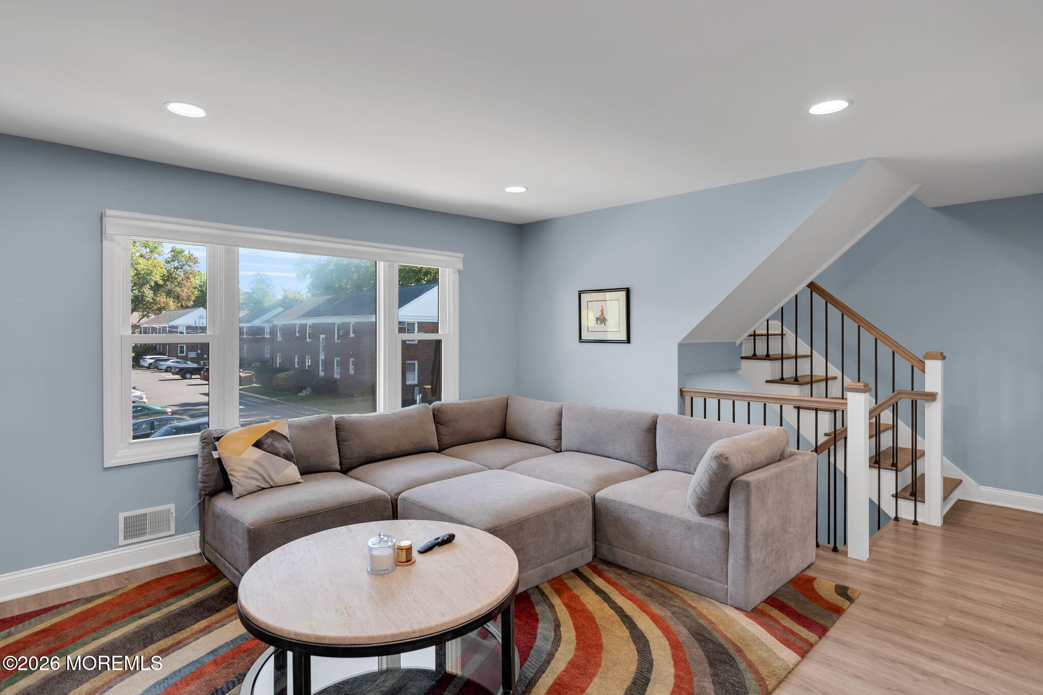 Living room with a gray sectional, colorful striped rug, and staircase beside large windows.