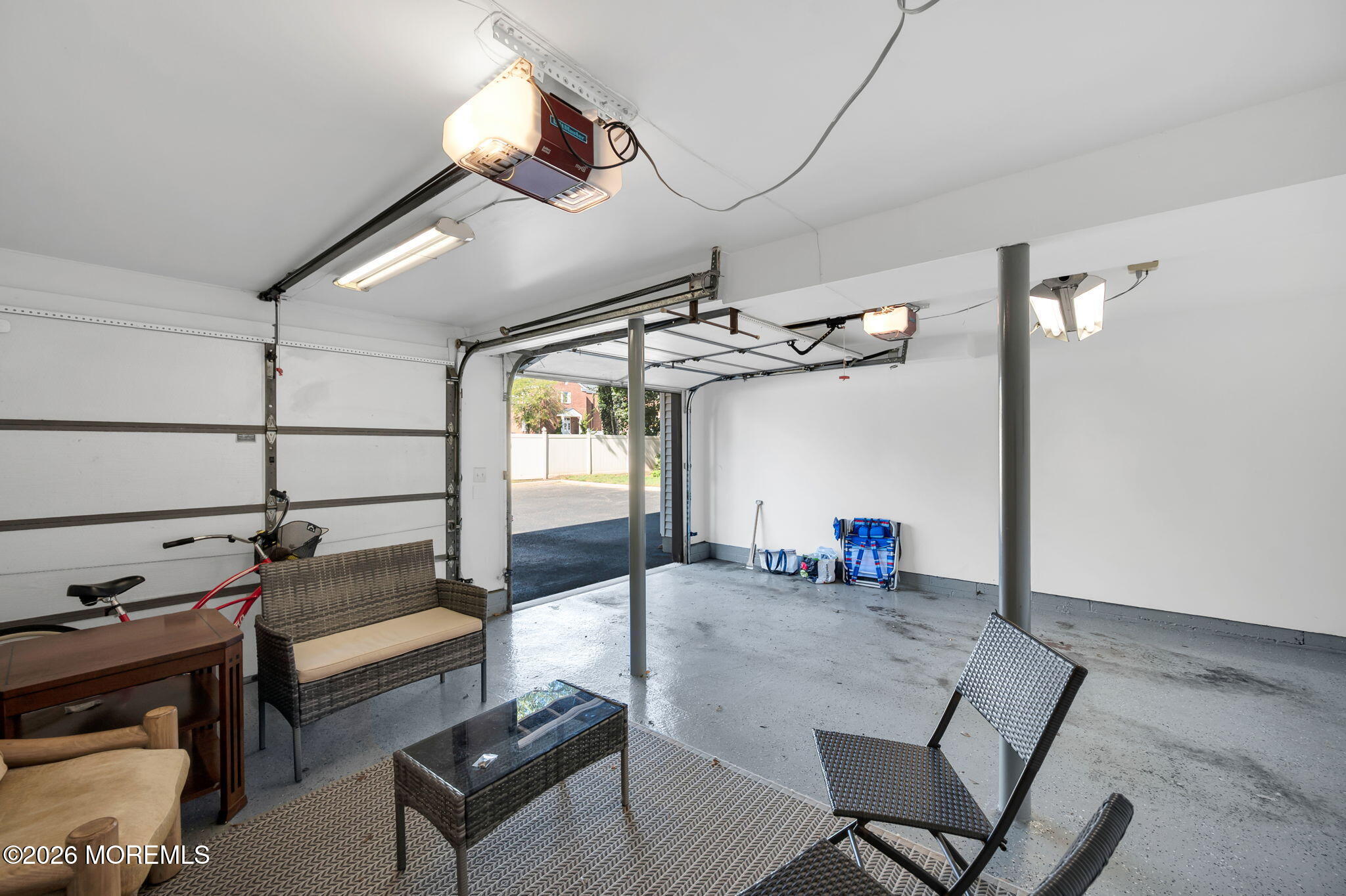 Garage with an open overhead door, concrete floor, and wicker seating on a rug.