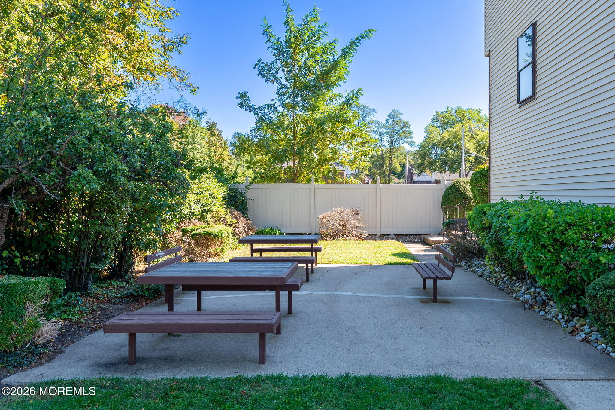 Outdoor patio with picnic tables and benches, bordered by shrubs and a white privacy fence.