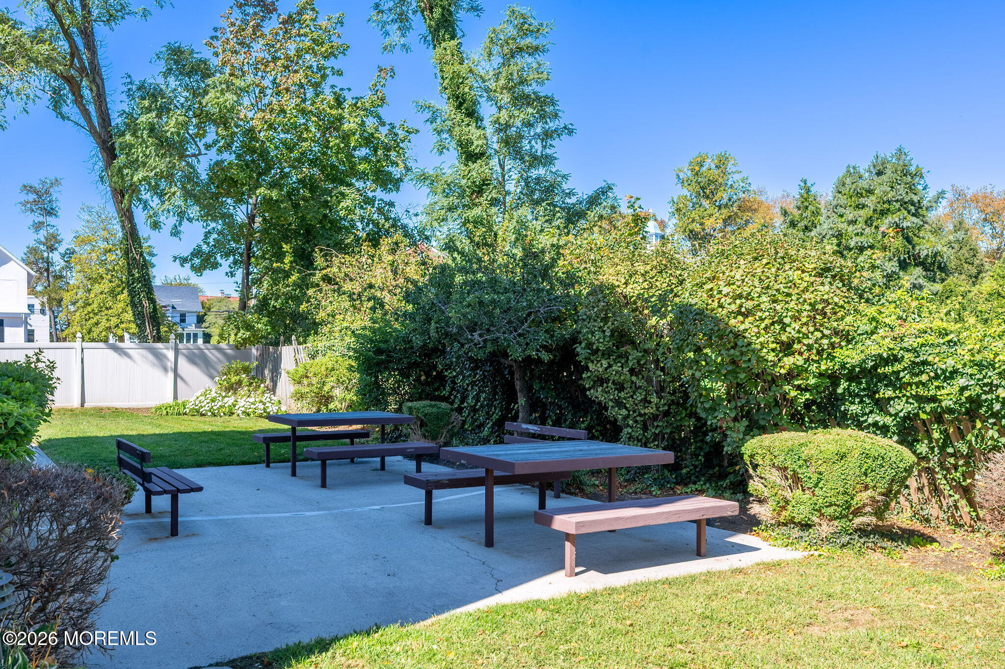 Outdoor patio with picnic tables and benches, surrounded by hedges and mature trees.