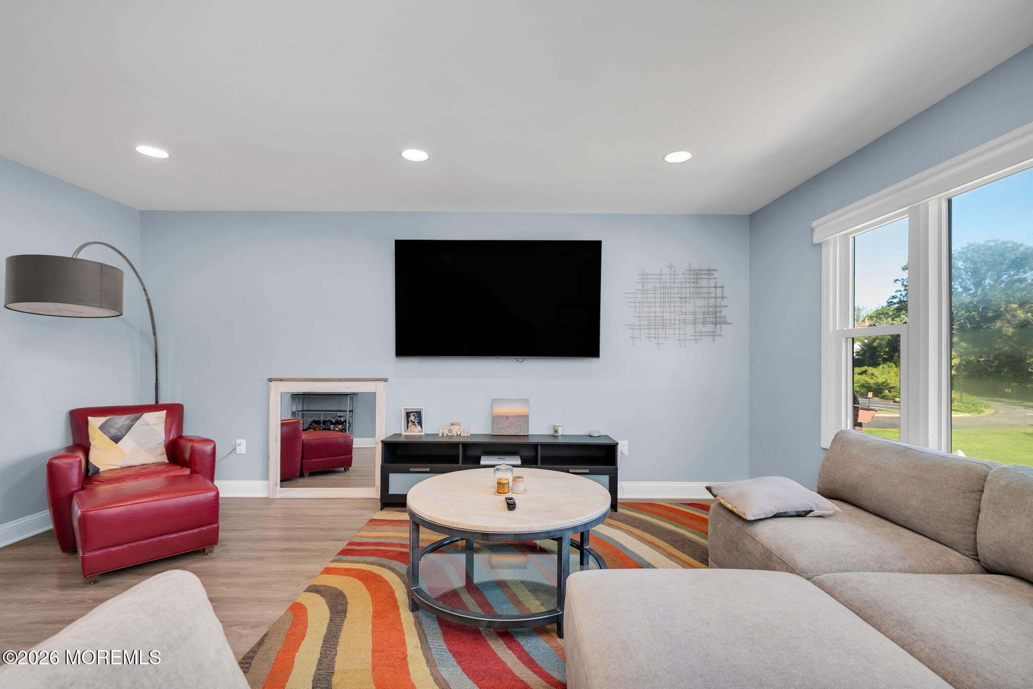 Furnished living room with a mirrored fireplace, red leather armchair, colorful rug, and large windows overlooking greenery.