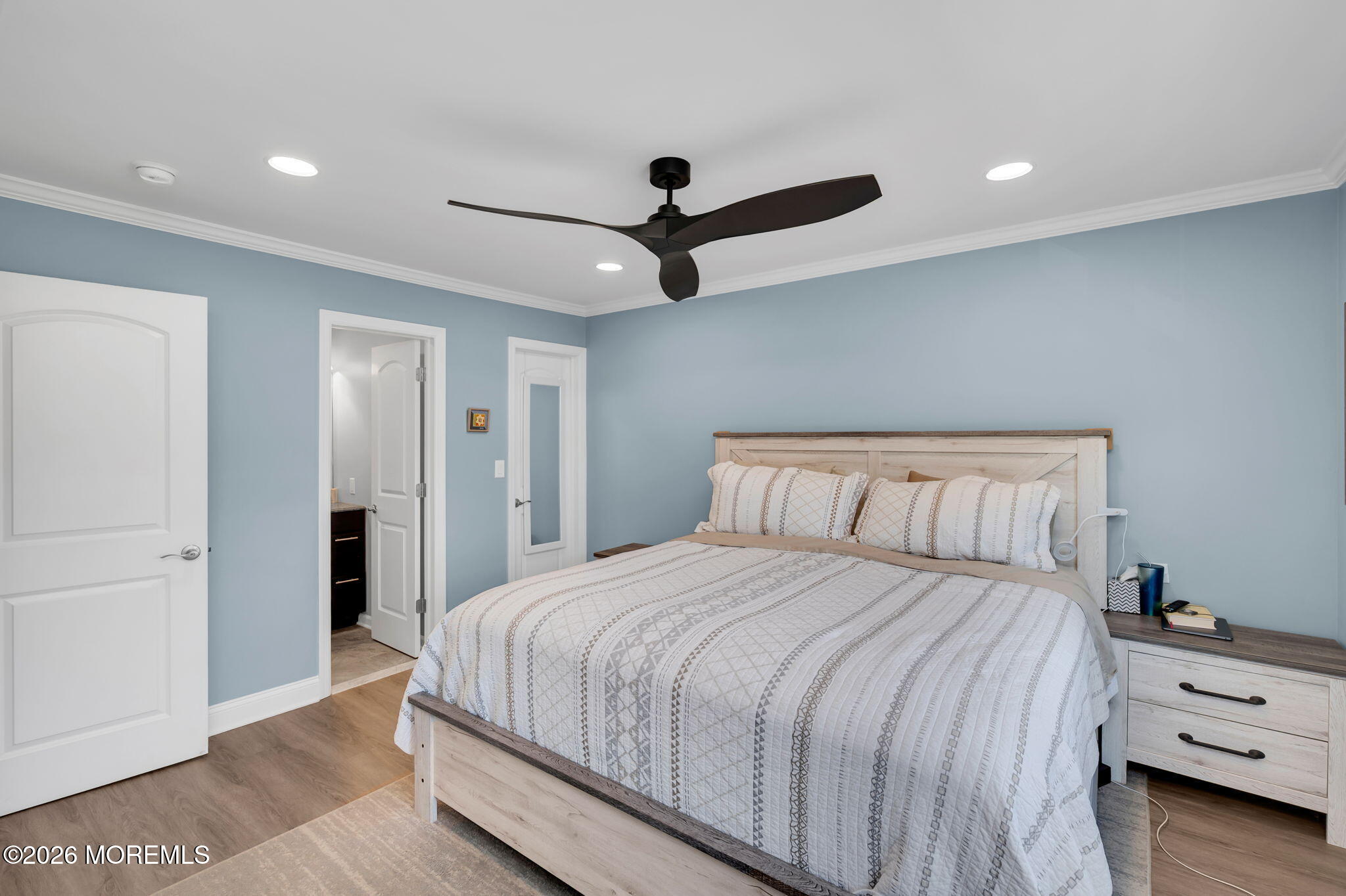 Bedroom with pale blue walls, patterned white bedding, light wood furniture, and a dark ceiling fan.
