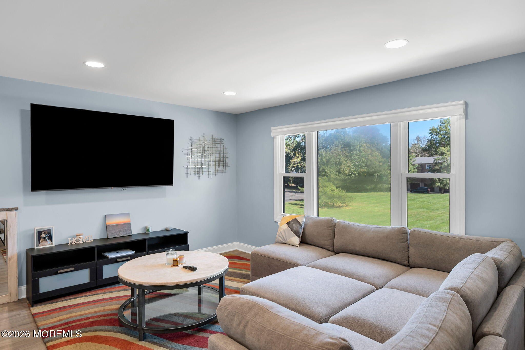 Living room with a large gray sectional sofa, wall-mounted television, and windows overlooking a green lawn.