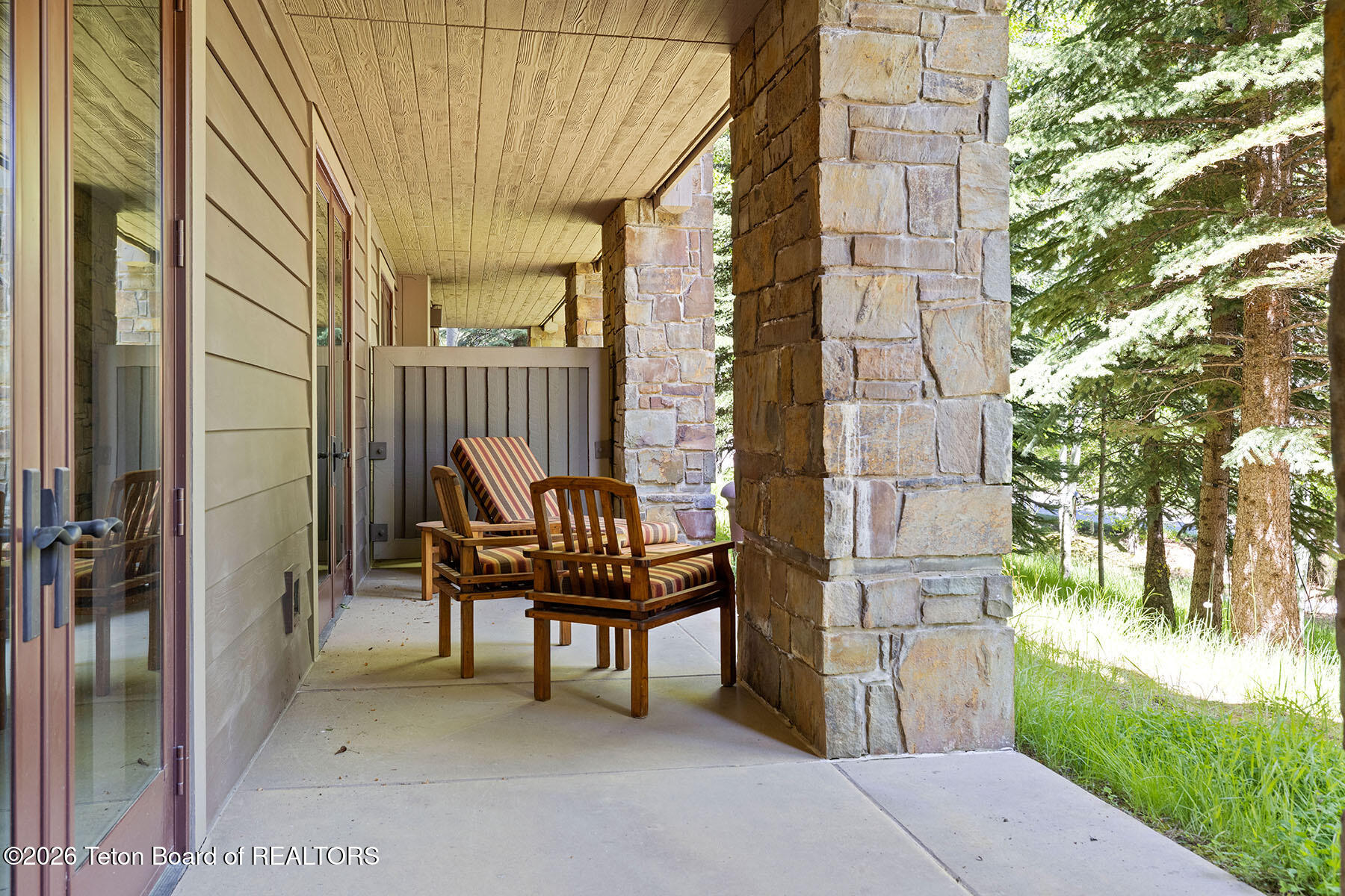 Covered porch with stone columns and striped-cushion chairs beside a wooded yard.