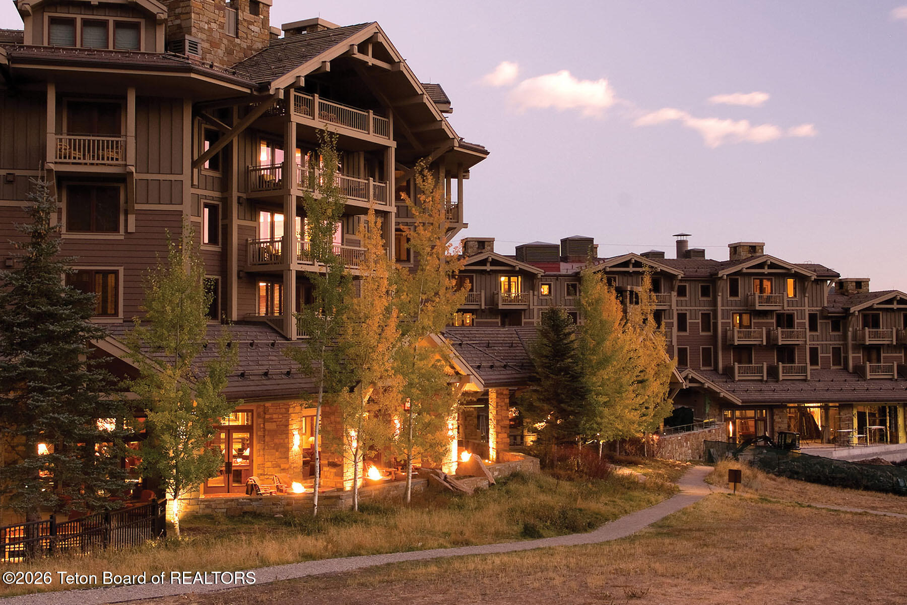 Mountain-style condominium buildings with balconies and glowing windows amid autumn trees at dusk.