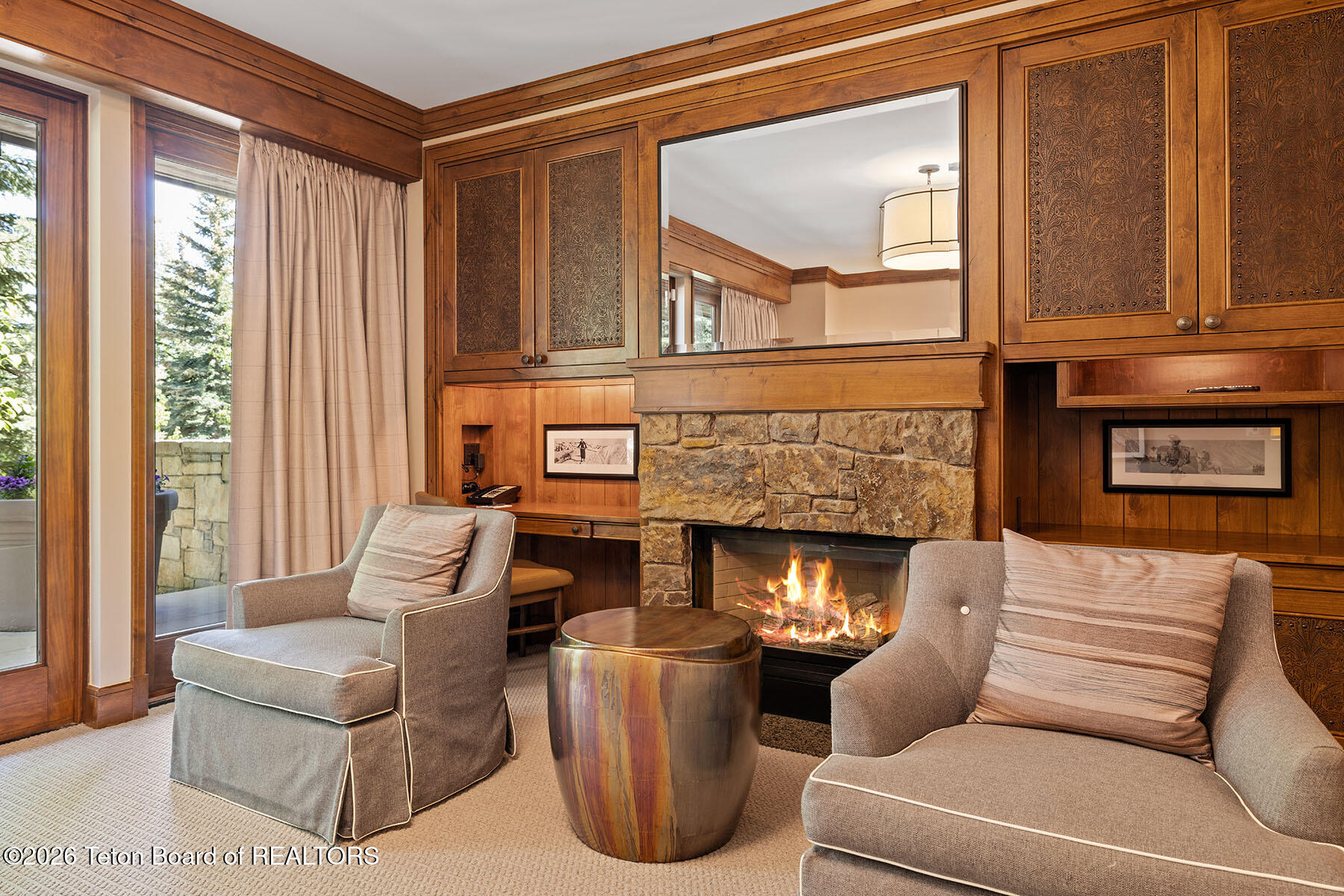 Living room with a lit stone fireplace, ornate wood cabinetry, and windows overlooking trees.