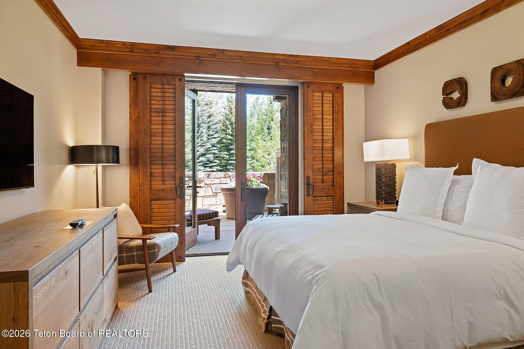Bedroom with a white bed and wood-trimmed doors opening onto a tree-lined patio.