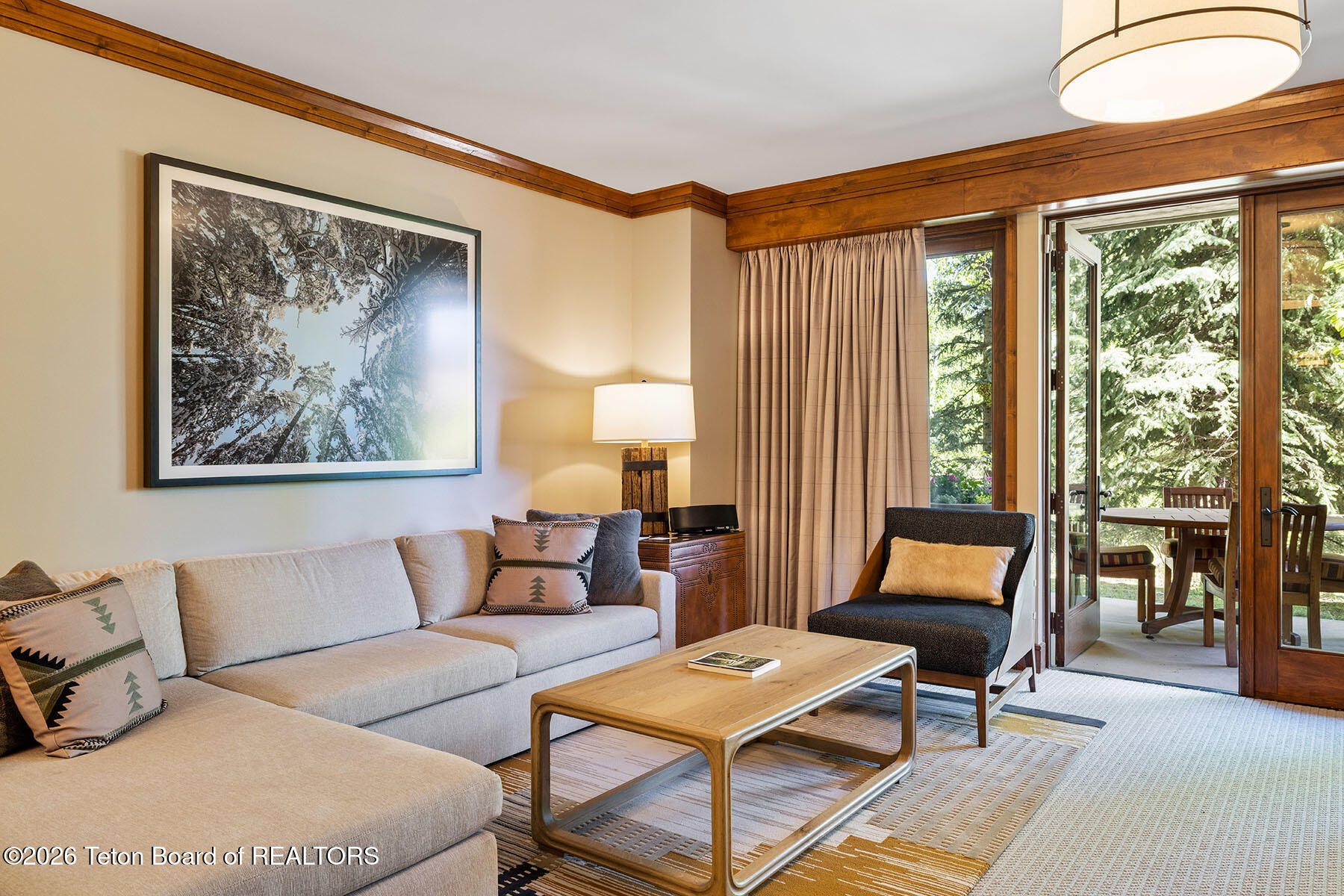 Living room with a pale sectional sofa and wood coffee table beside open doors to a tree-lined patio.