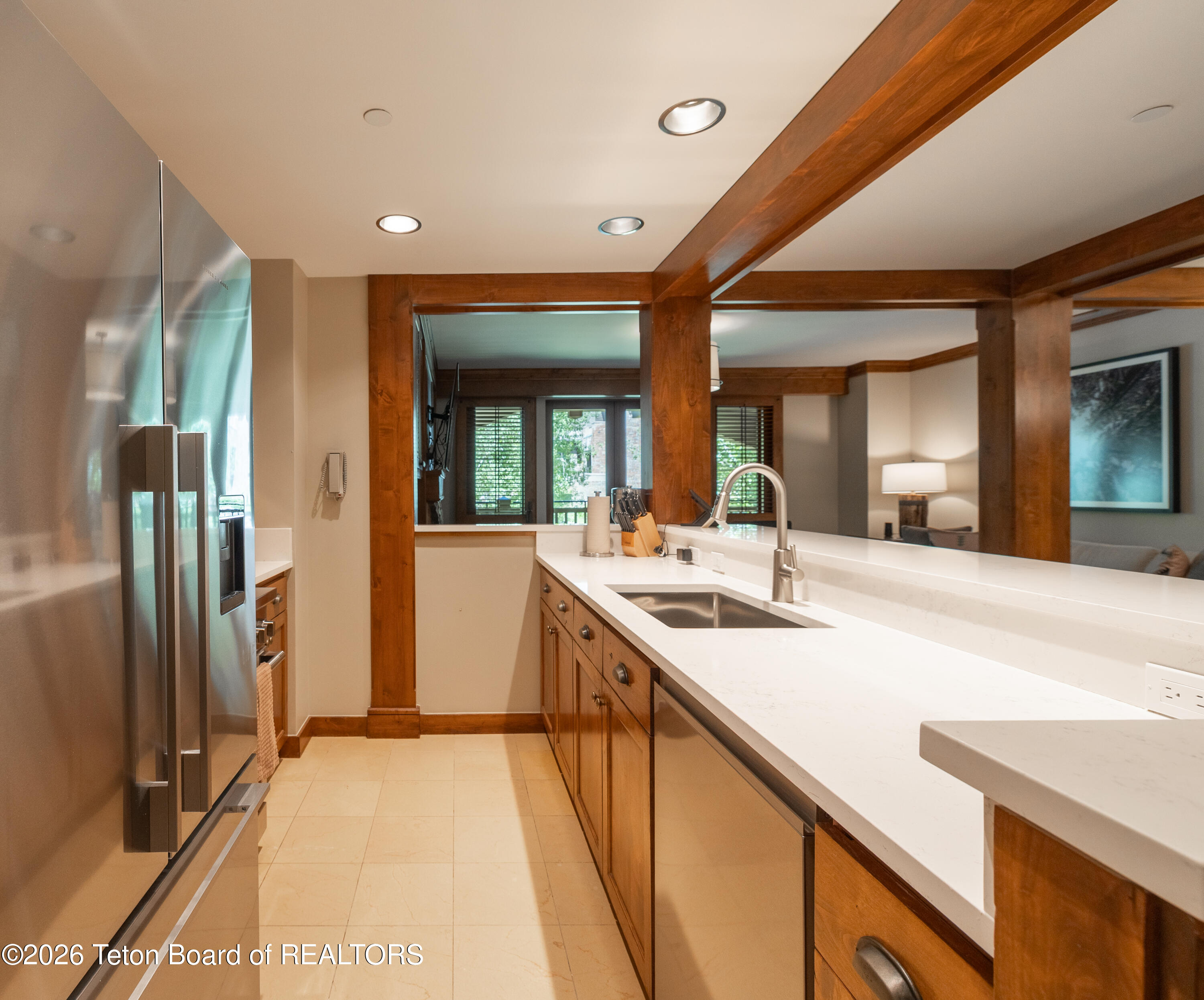 Kitchen with wood cabinetry, white counters, stainless refrigerator, and sink beneath wood ceiling beams.