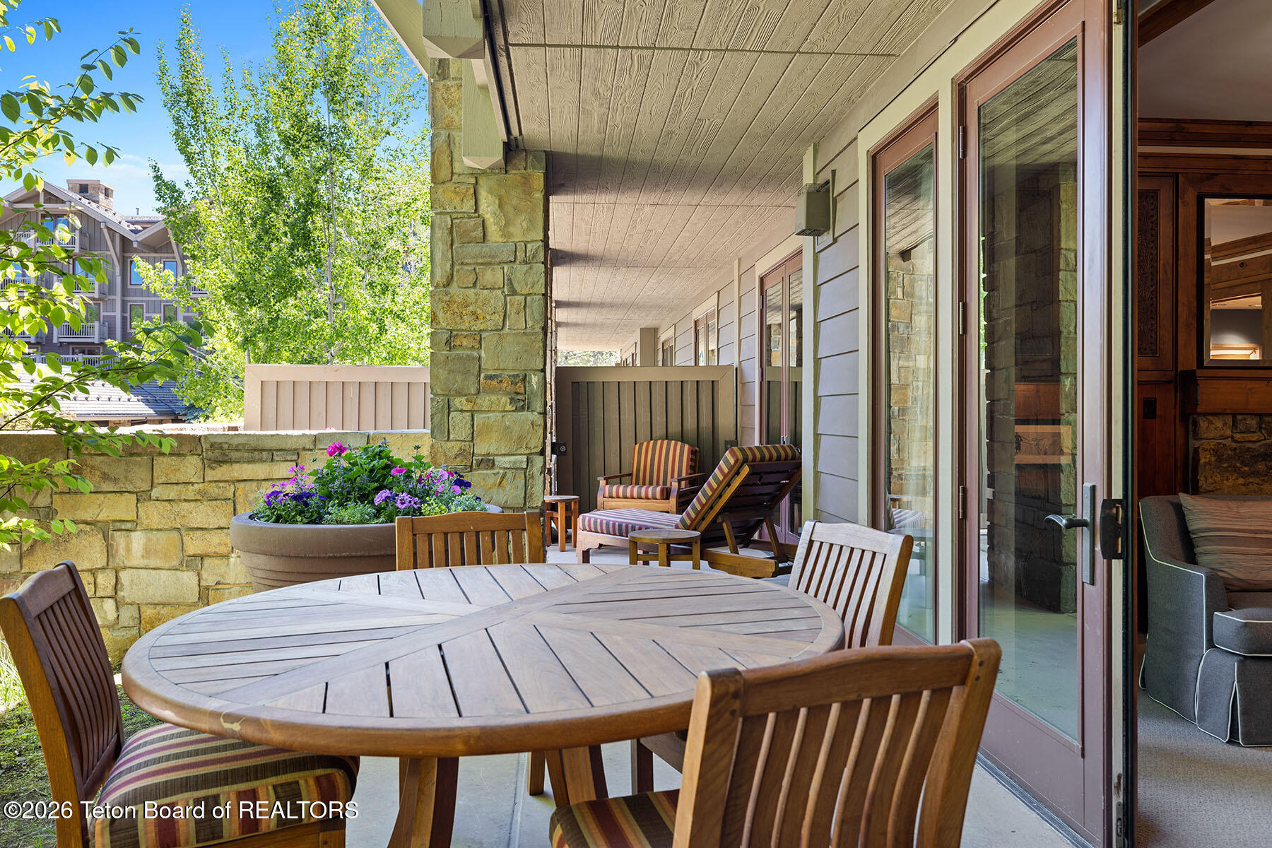 Covered patio with a round wooden dining table, striped lounge chairs, and a flower-filled planter.