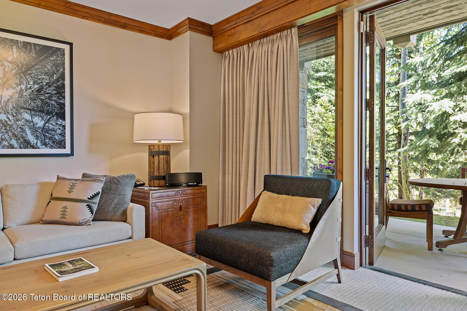 Living room with a light sofa and dark lounge chair beside an open patio door overlooking trees.