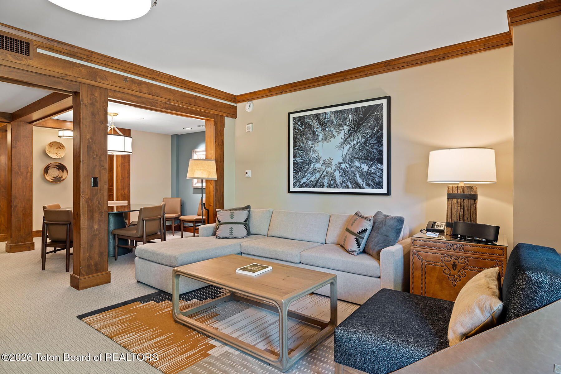 Living room with a gray sectional, patterned rug, and prominent timber beams framing the dining area.