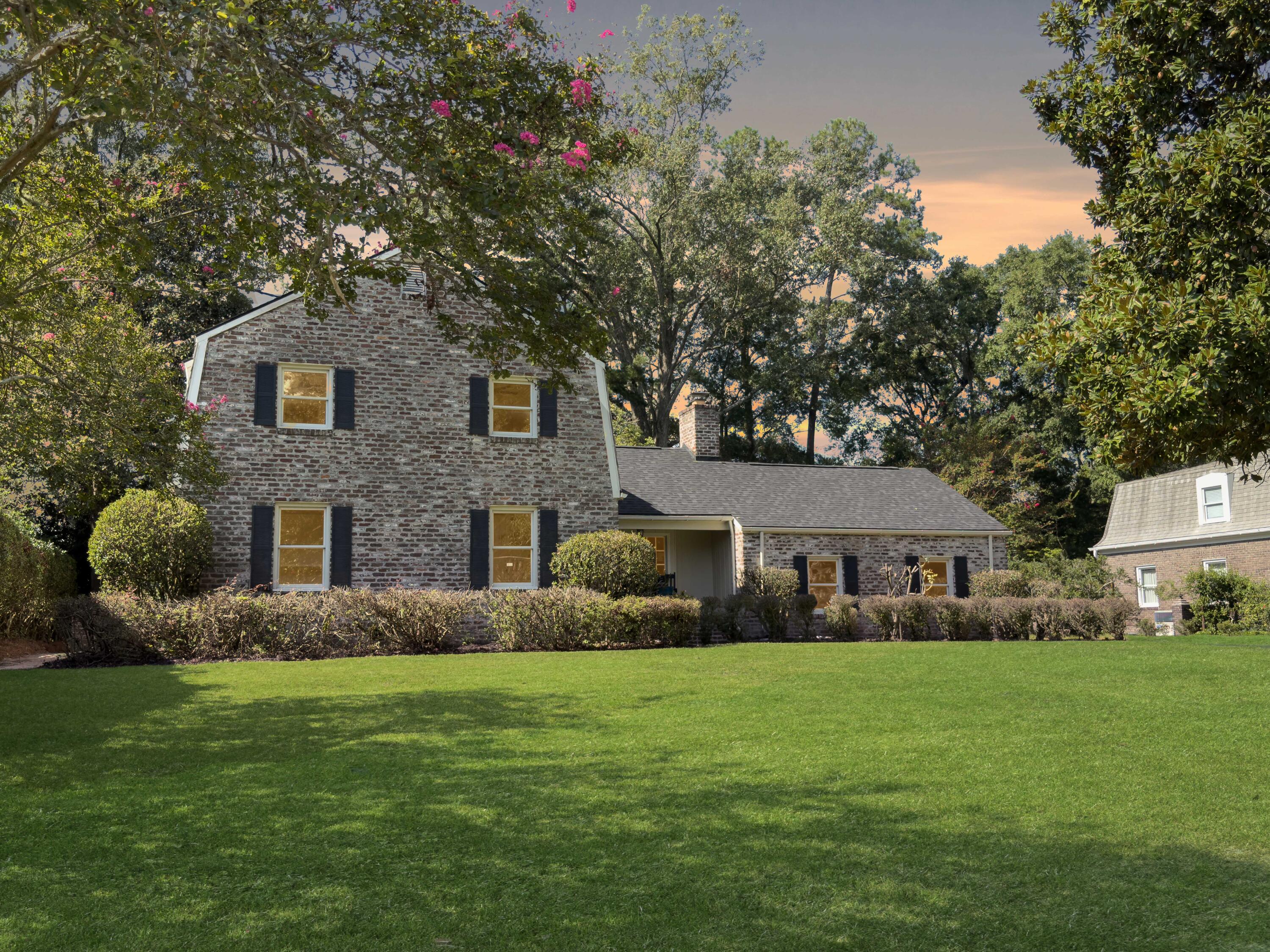 Two-story brick home with black shutters, a broad lawn, and flowering trees in warm evening light.