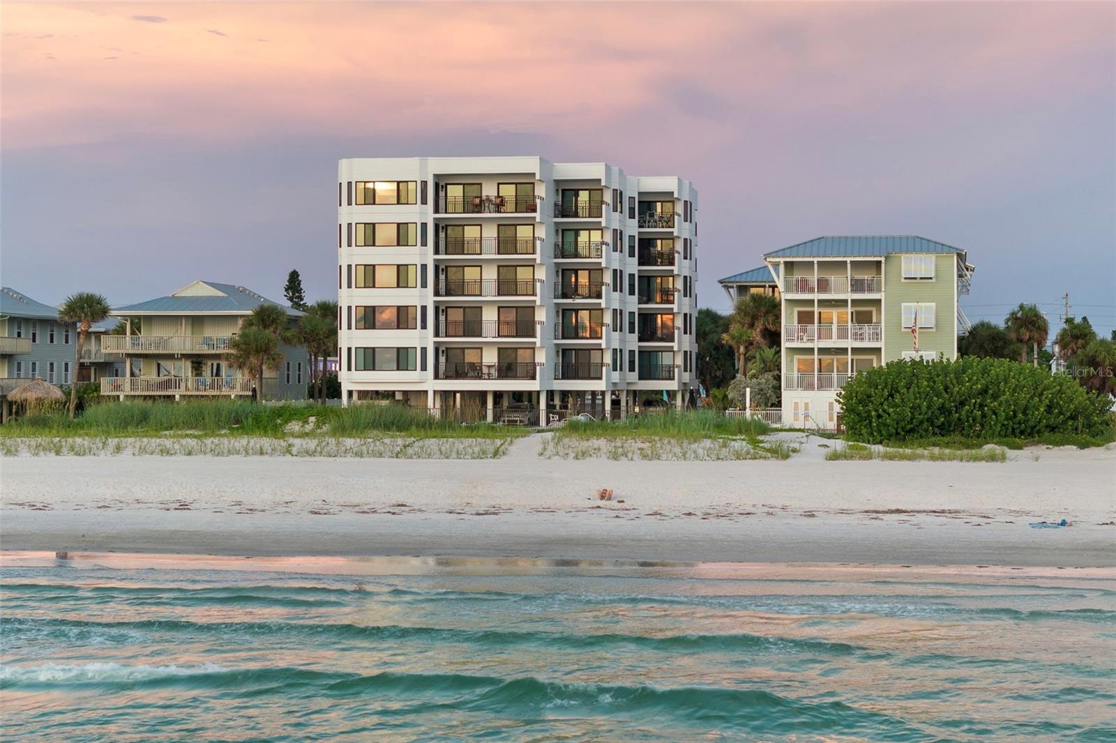 Beachfront condominium and neighboring homes above the shore at sunset, with waves in the foreground.