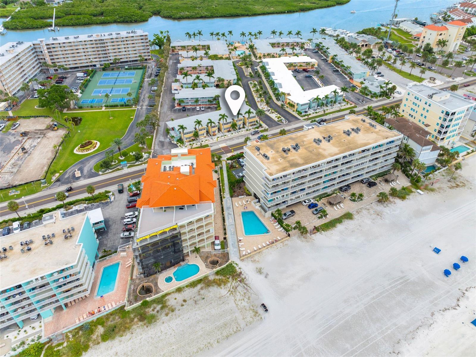 Aerial view of the beachfront condominium marked by a pin, with pools, tennis courts, and a waterway nearby.