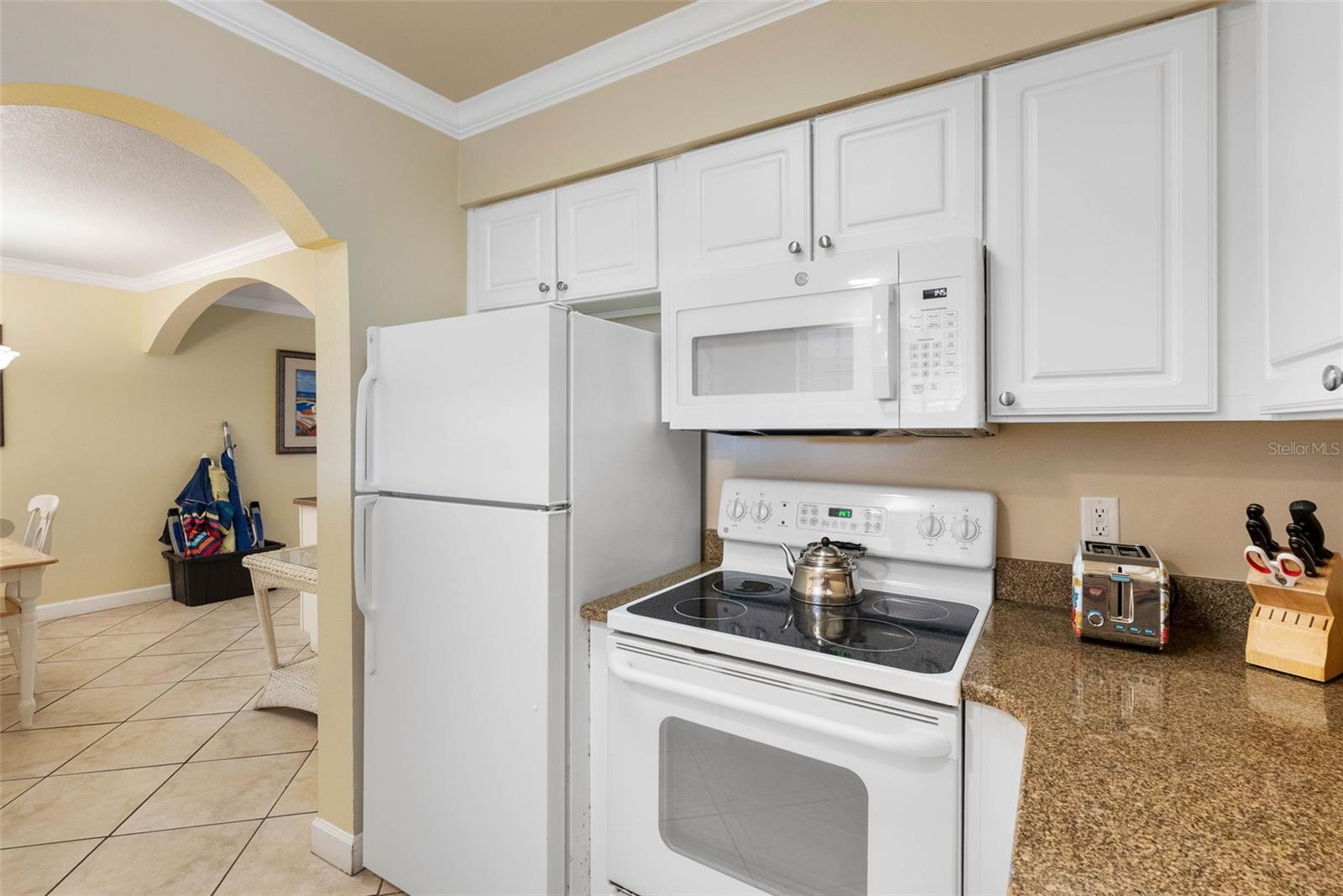 Kitchen with white cabinets, granite countertops, and white appliances, opening to a tiled dining area.
