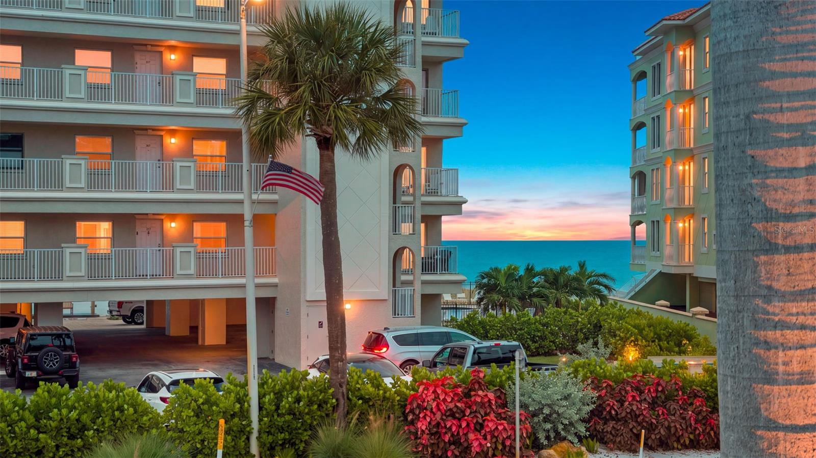 Condominium buildings overlook the Gulf at sunset, with a palm tree and American flag.