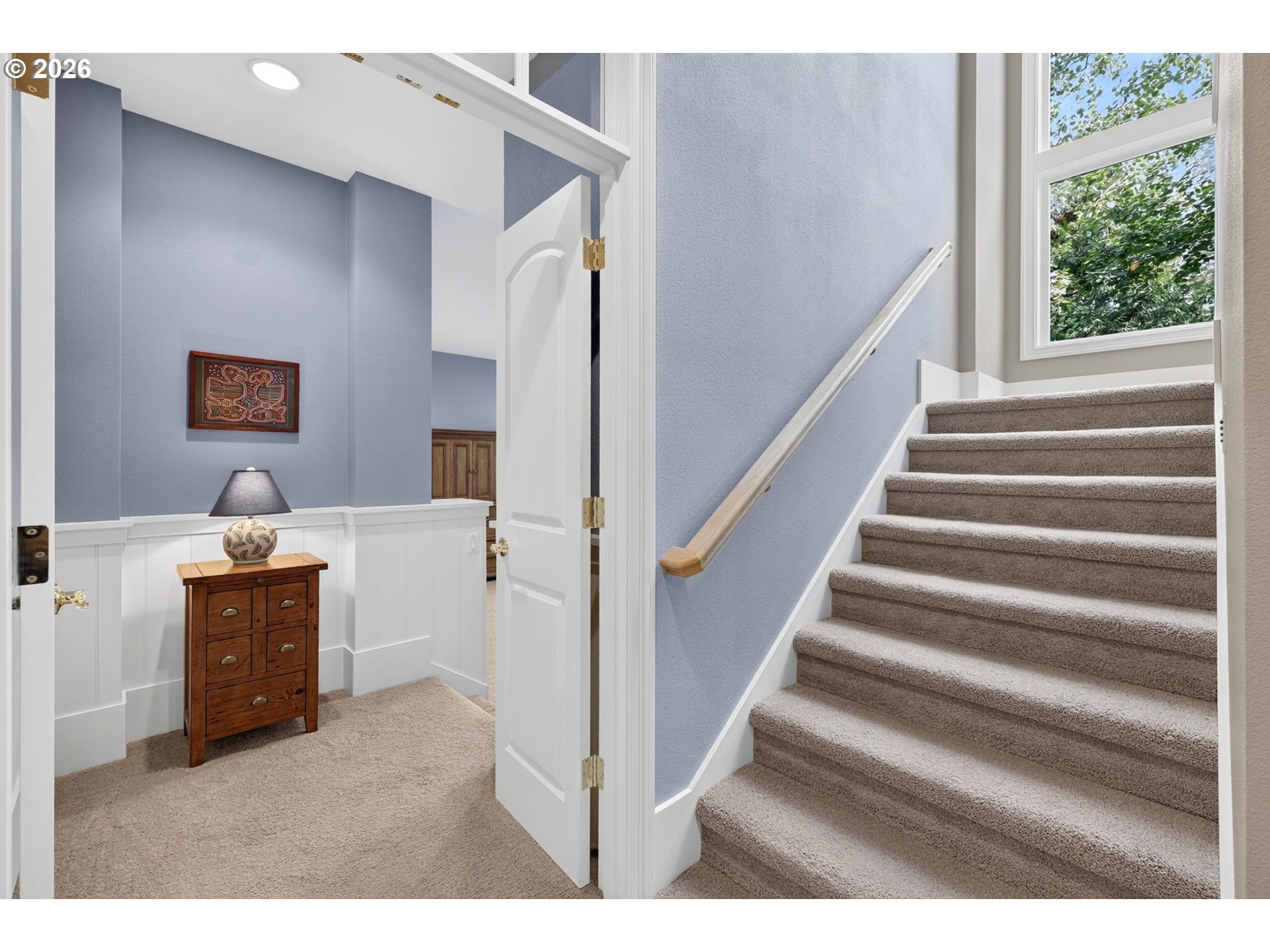 Carpeted staircase beside a landing with a wooden chest, lamp, and window overlooking trees.