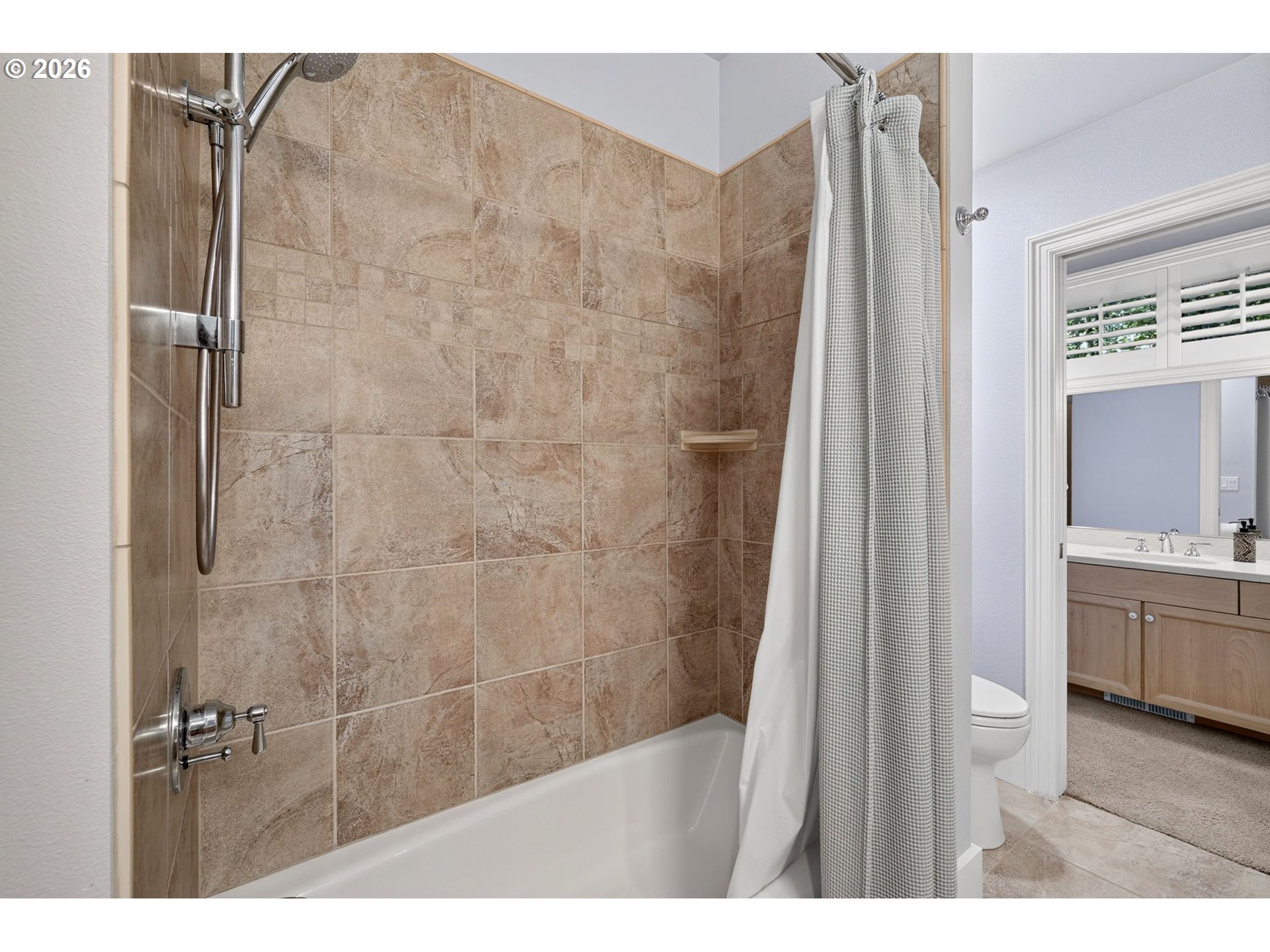 Bathroom tub-shower with tan tiled walls and an adjoining vanity and toilet.