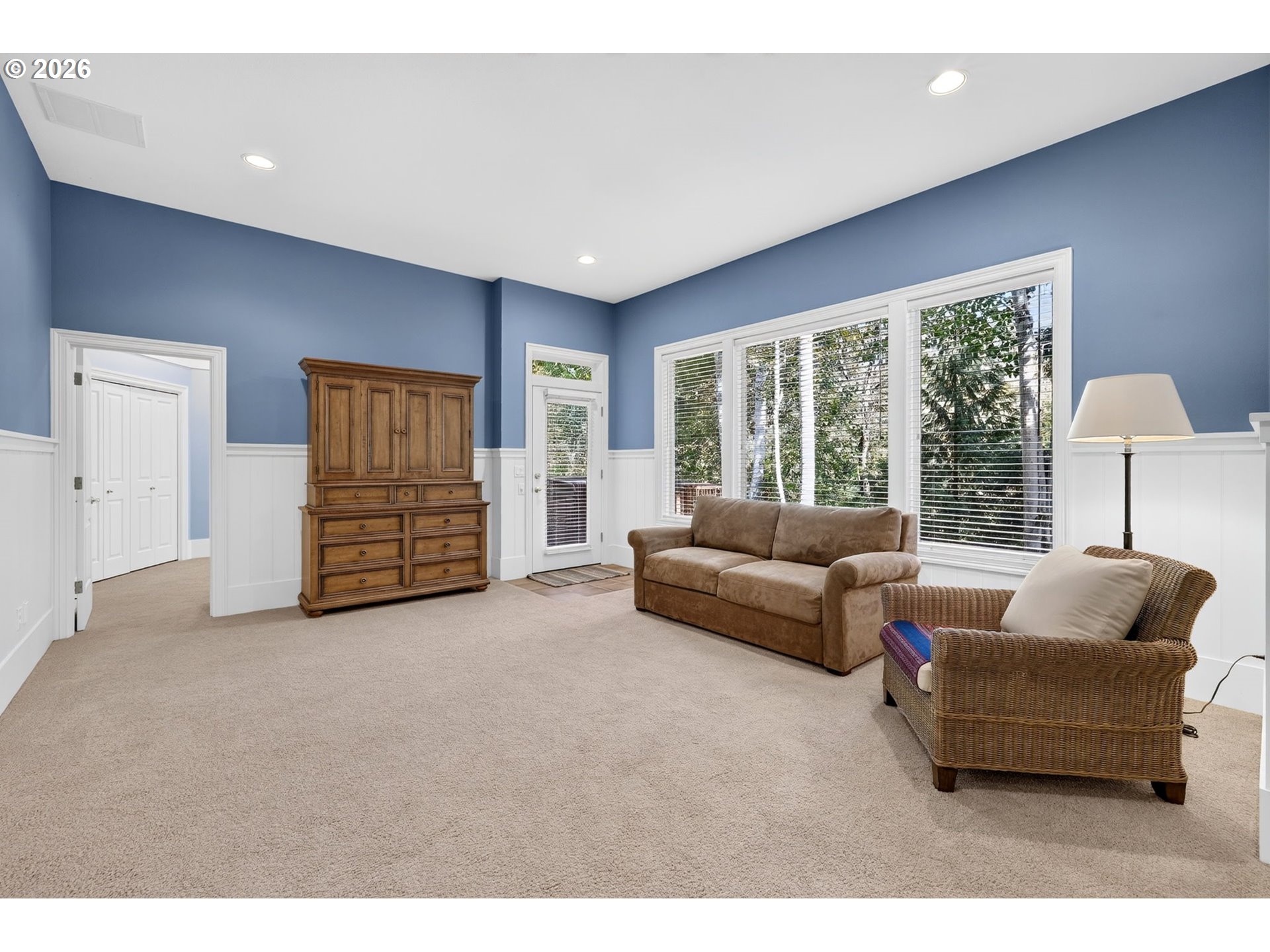 Living room with blue walls, a brown sofa, wicker chair, and large windows overlooking trees.