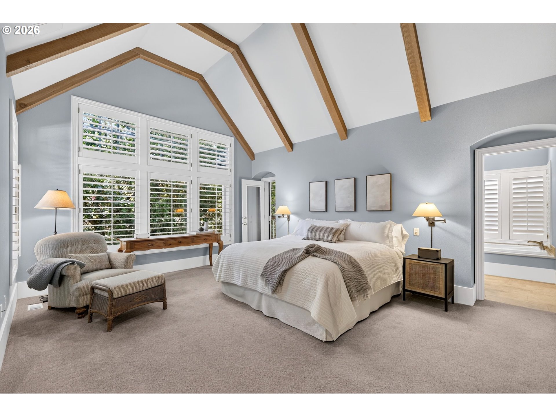 Vaulted bedroom with exposed wood beams, shuttered windows, a large bed, and a cushioned sitting area.