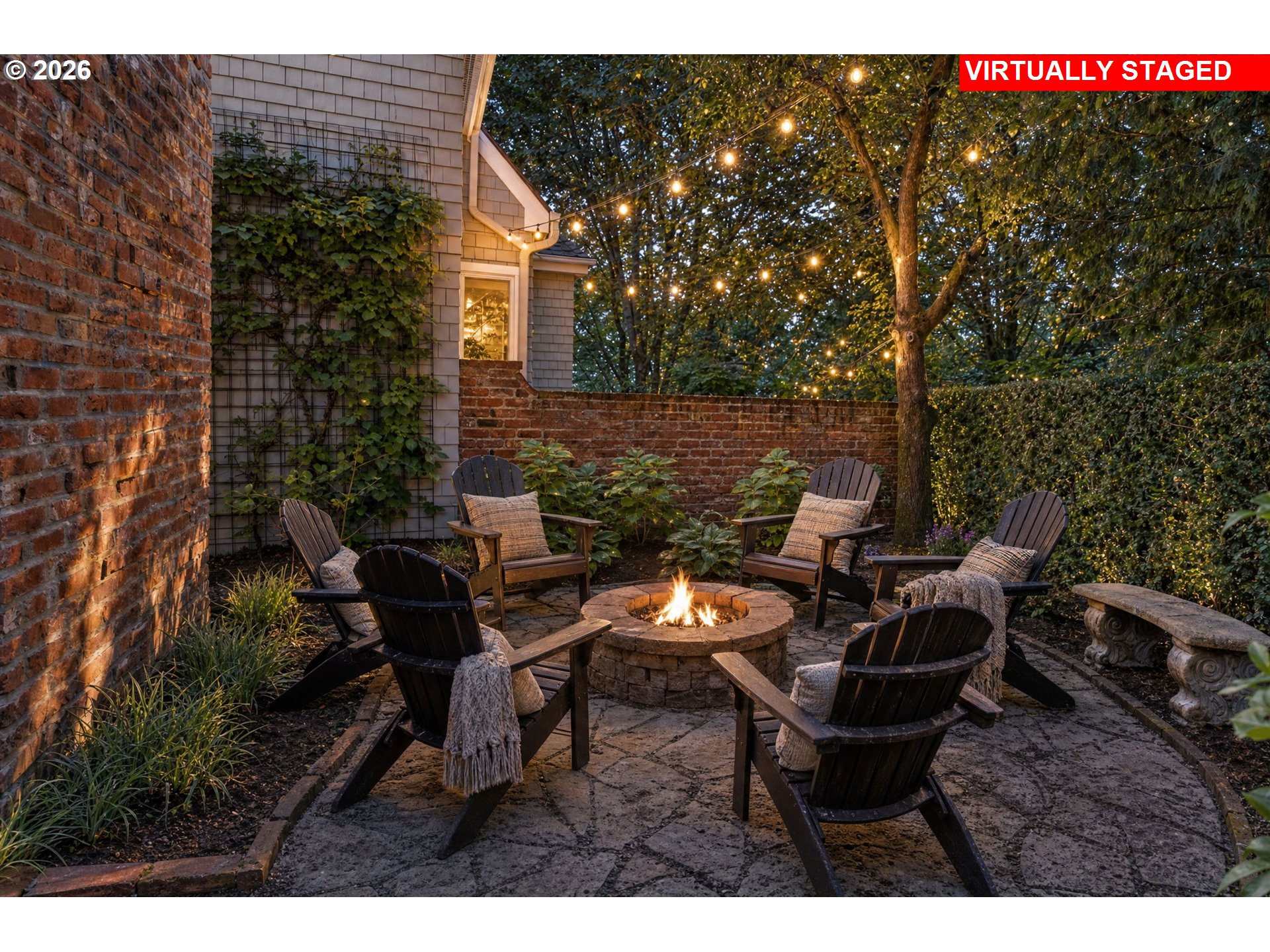 Backyard patio with Adirondack chairs around a stone fire pit beneath glowing string lights.