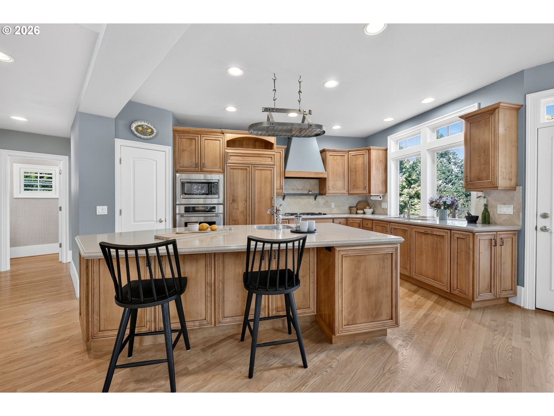 Kitchen with warm wood cabinetry, a large island, black bar stools, and wide windows overlooking greenery.