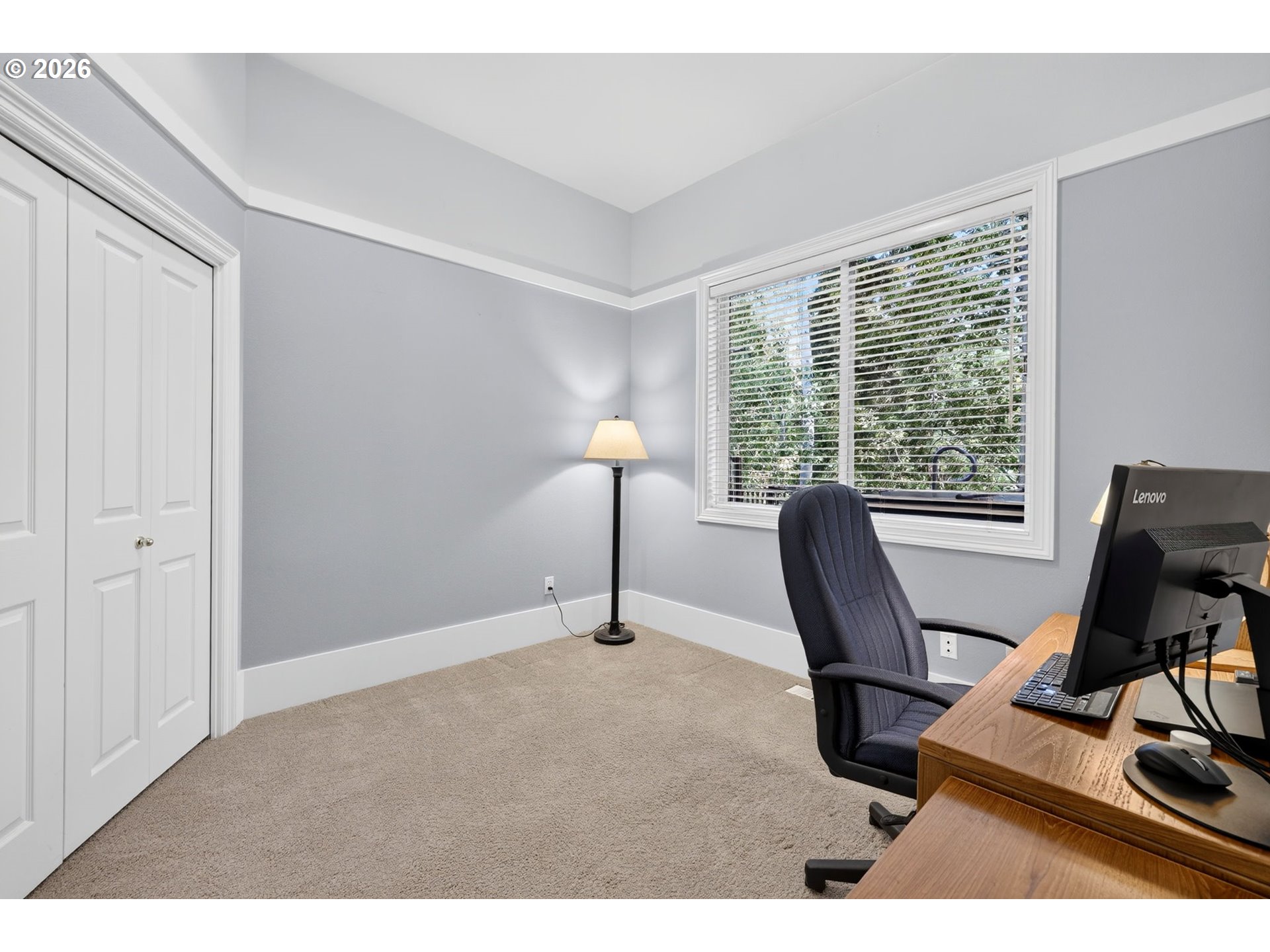Home office with a wooden computer desk, dark chair, closet doors, and a large window overlooking trees.