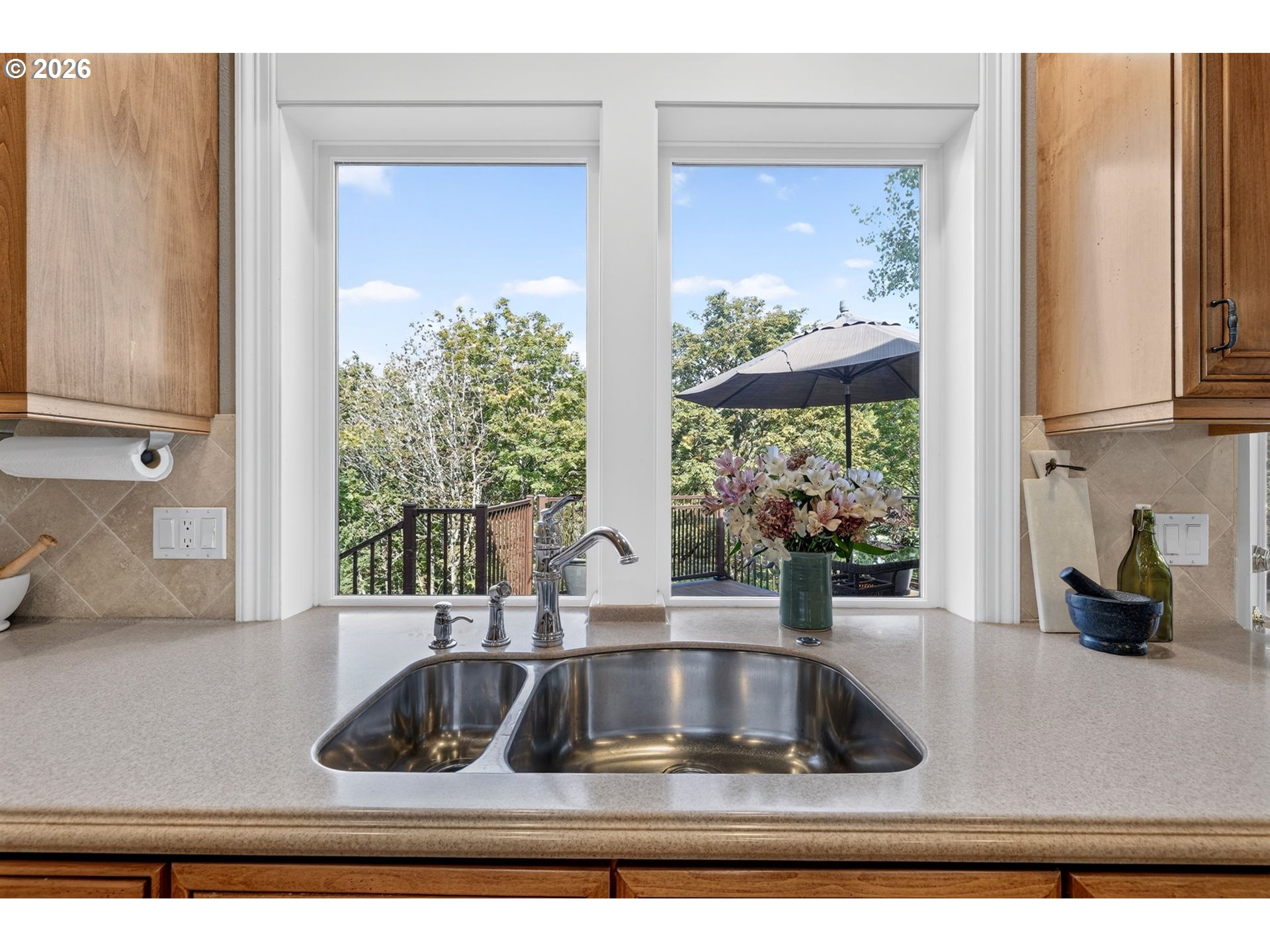 Kitchen sink beneath a double window overlooking a deck and leafy trees.