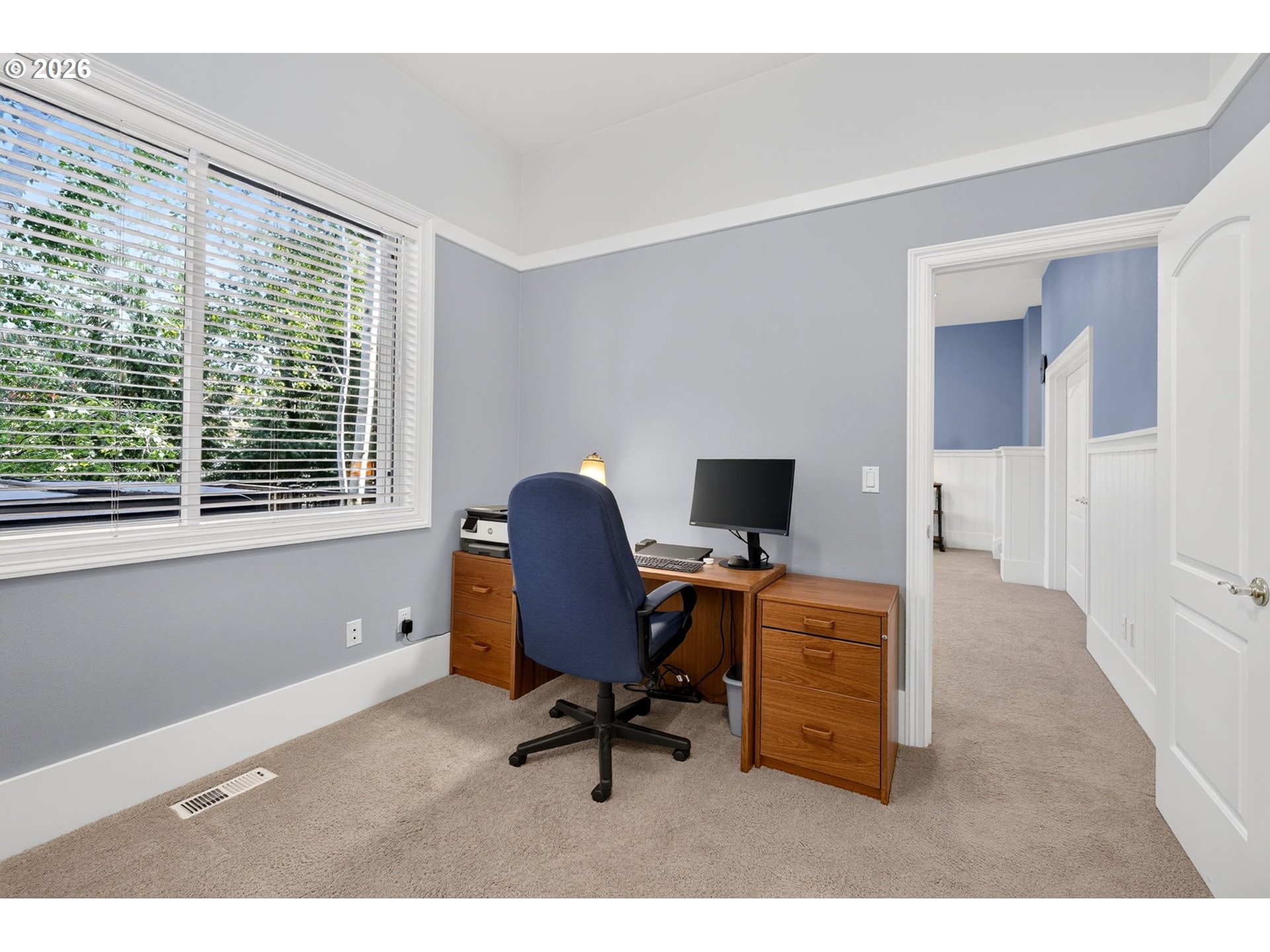 Home office with a wooden desk and blue chair beside a large window with white blinds.