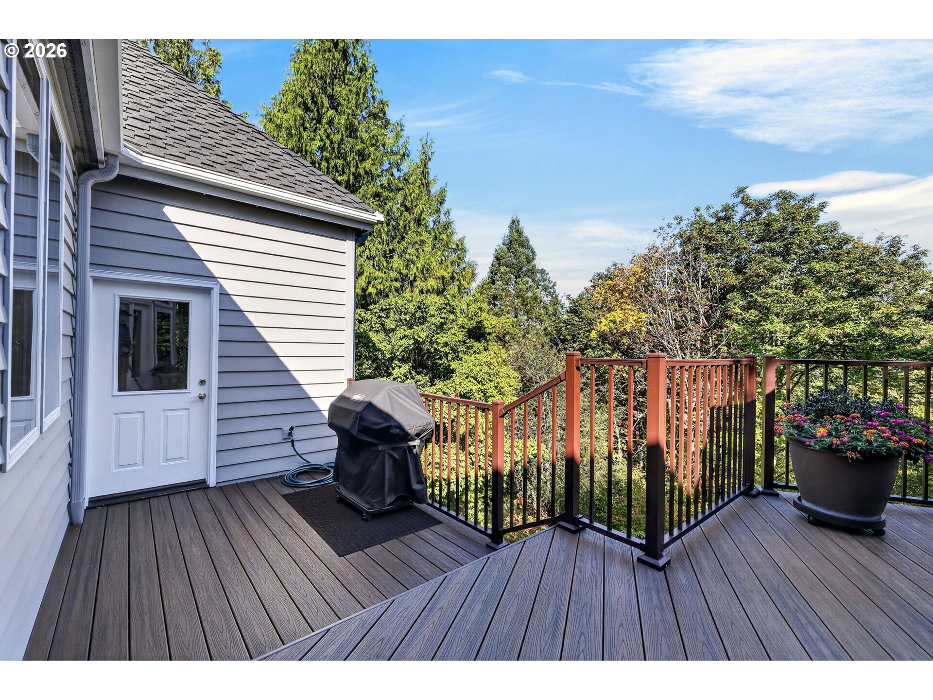 Raised deck beside a gray-sided home, with wood-toned railings, a covered grill, and a flower planter.