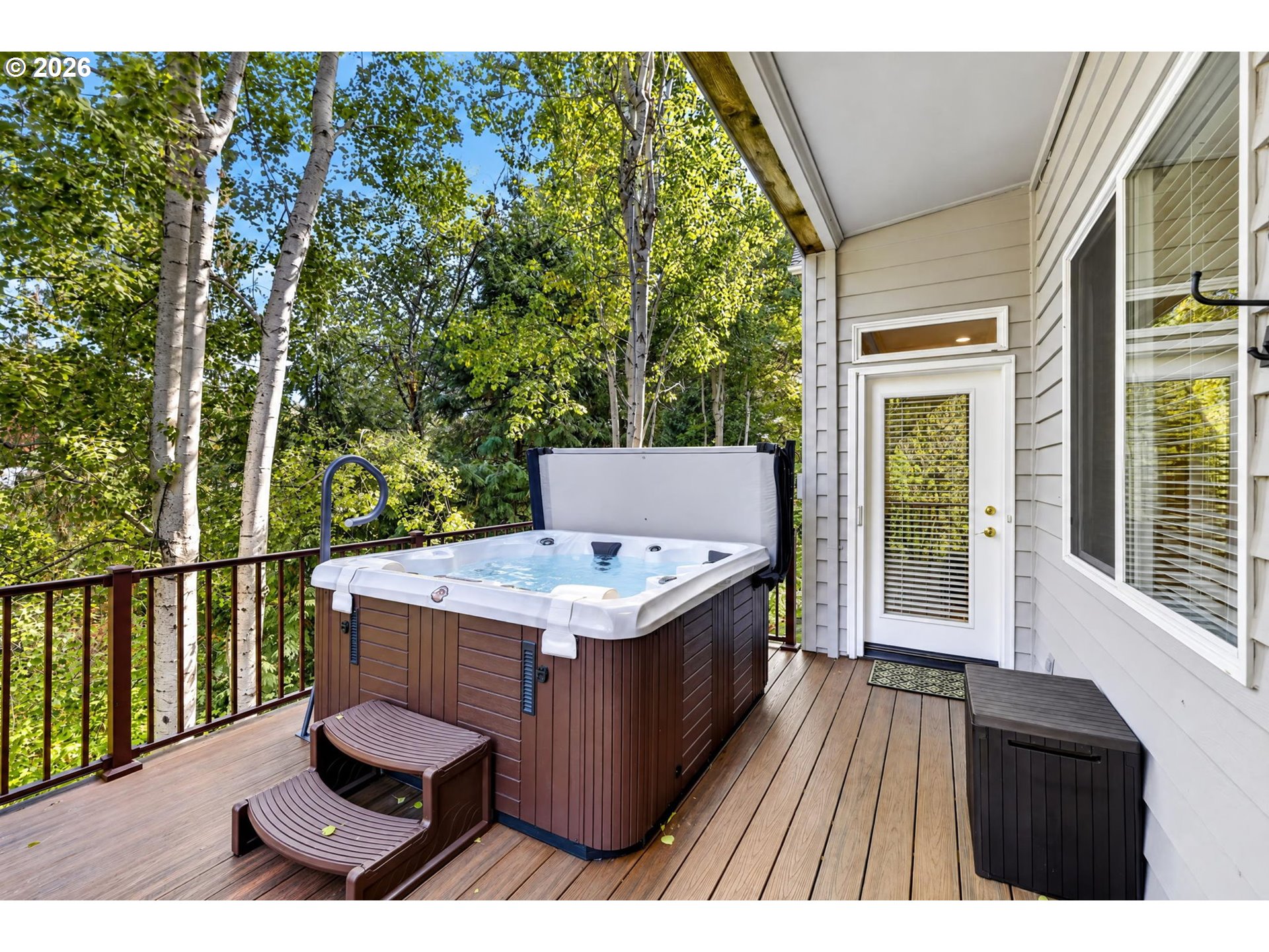 Deck with a large hot tub, entry steps, and leafy trees beyond the railing.
