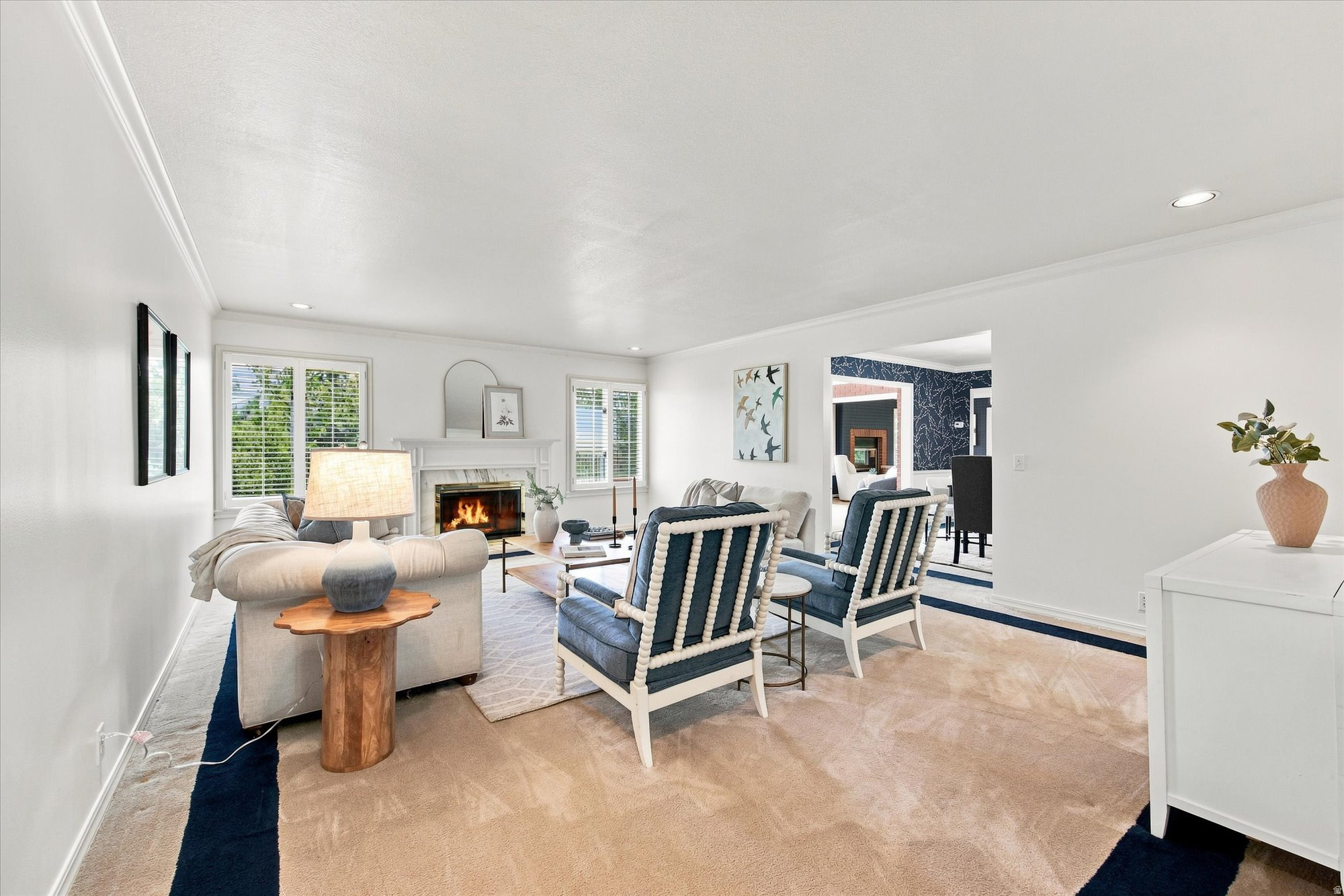 Staged living room with a lit fireplace, shuttered windows, and blue-and-white armchairs.