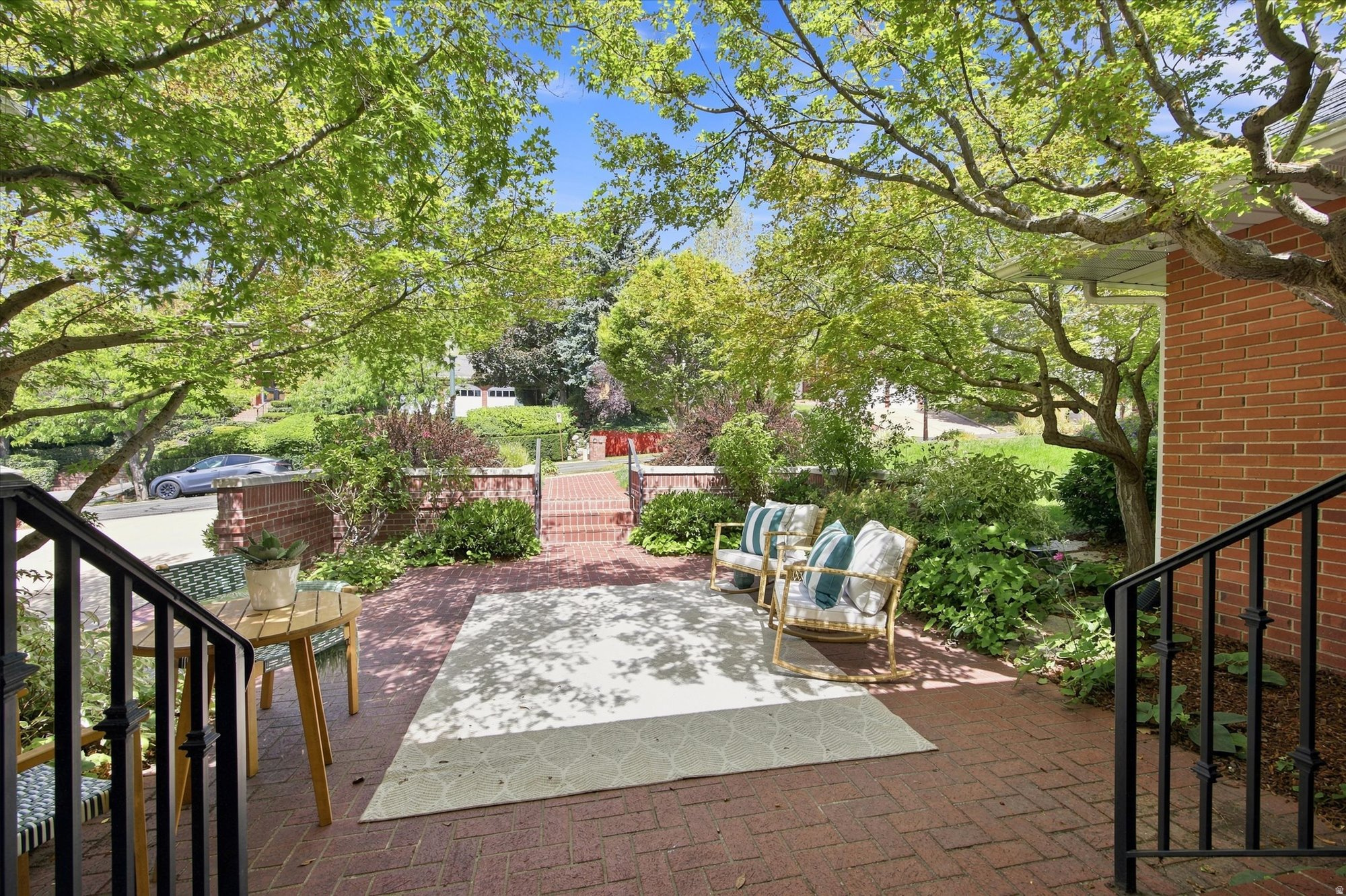 Shaded brick patio with cushioned chairs and a garden beneath mature trees.