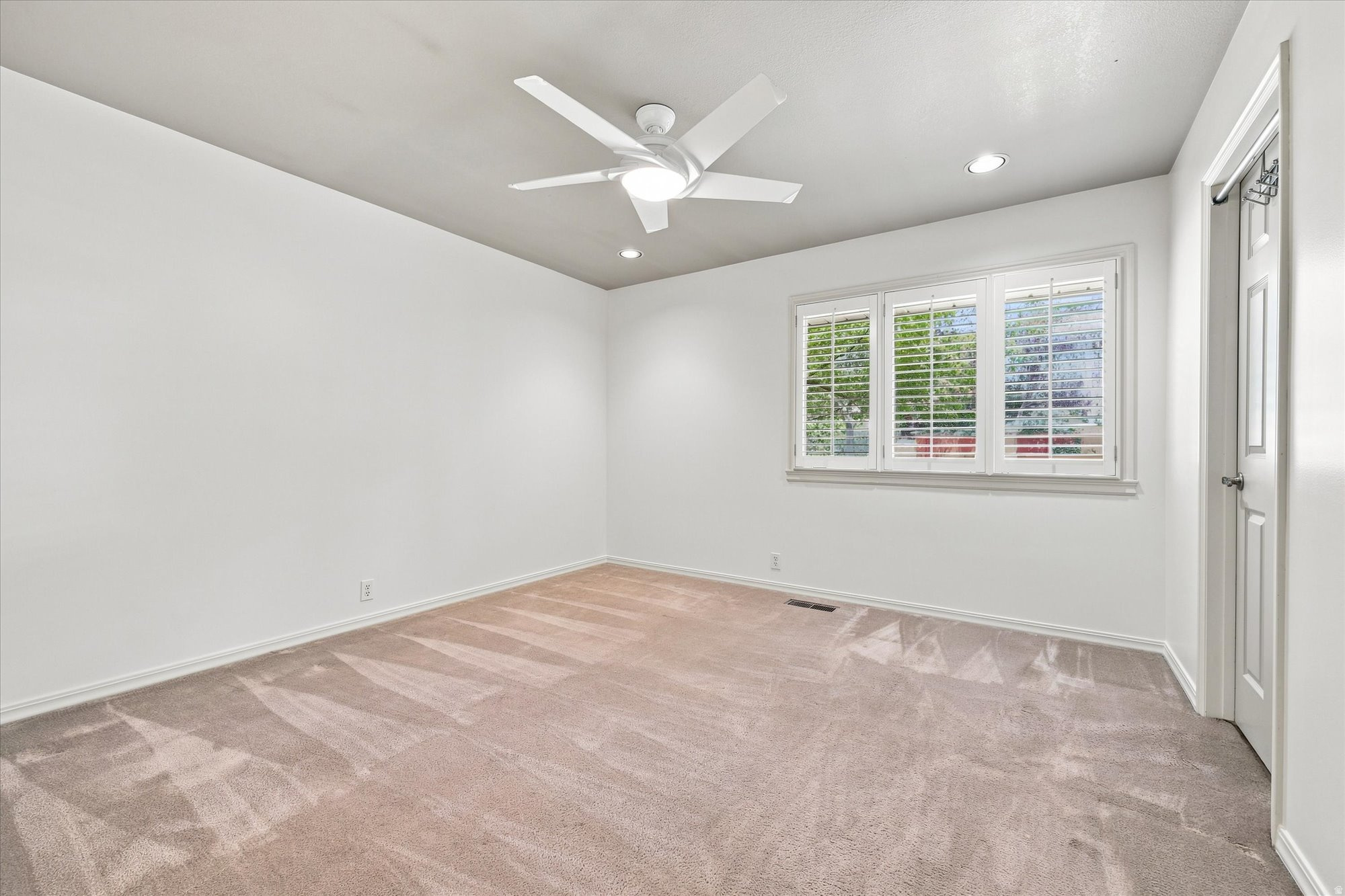 Empty carpeted bedroom with a ceiling fan and a wide plantation-shuttered window.