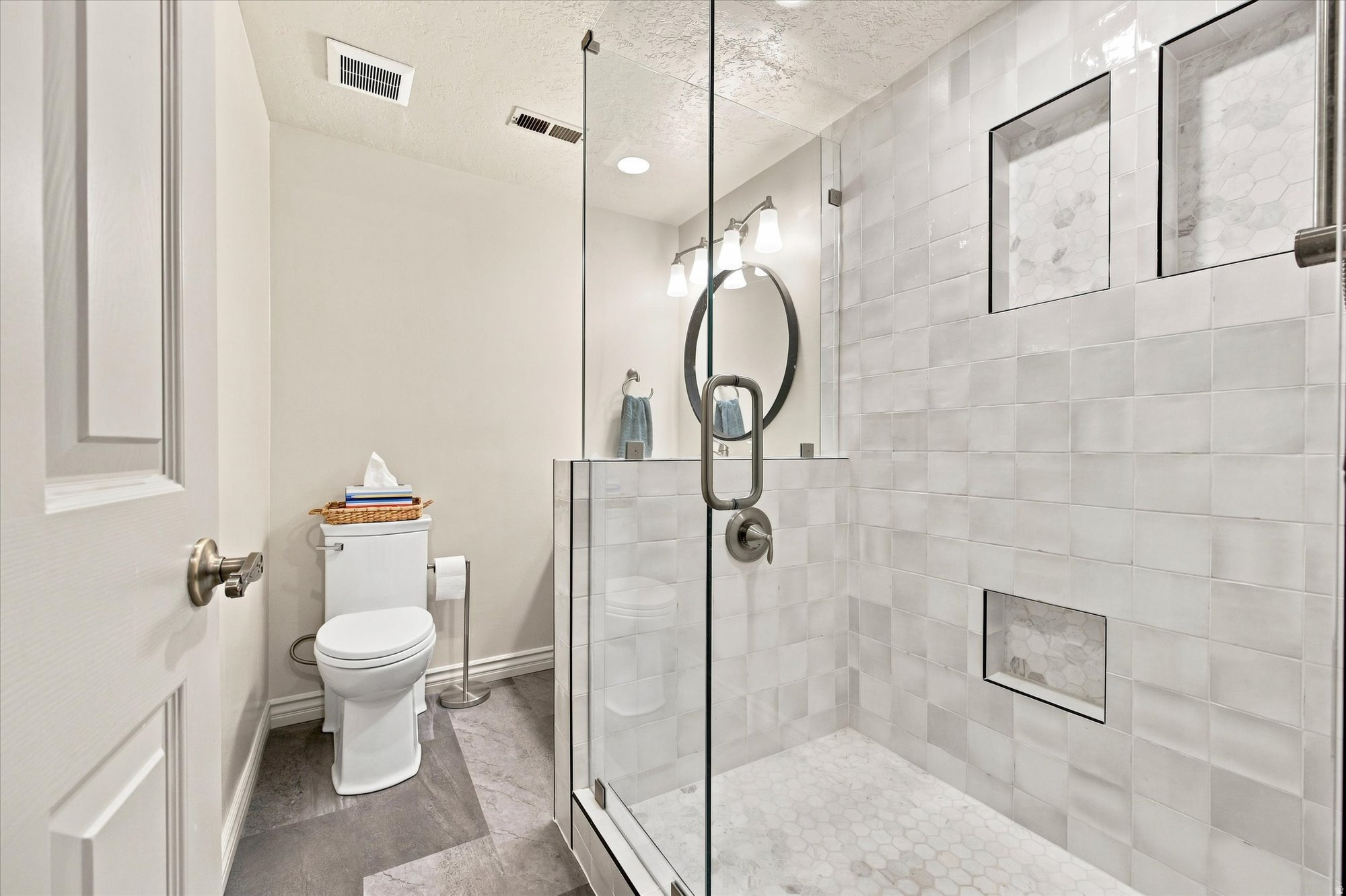 Bathroom with a glass-enclosed tiled shower, recessed niche, and white toilet beneath an oval mirror.