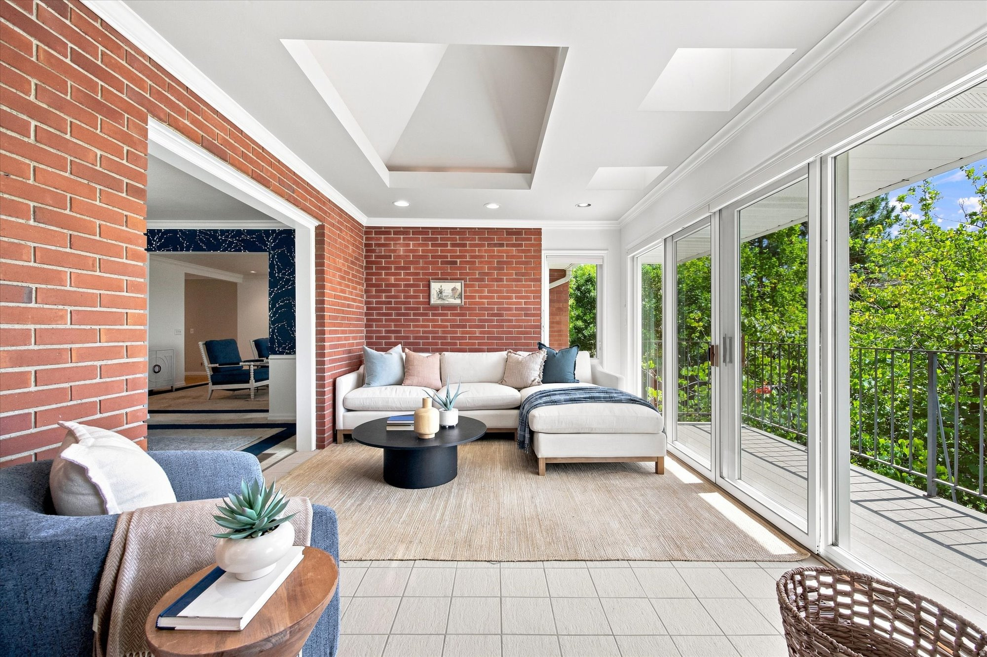 Furnished sunroom with exposed brick walls, a white sectional, and sliding glass doors to a leafy balcony.