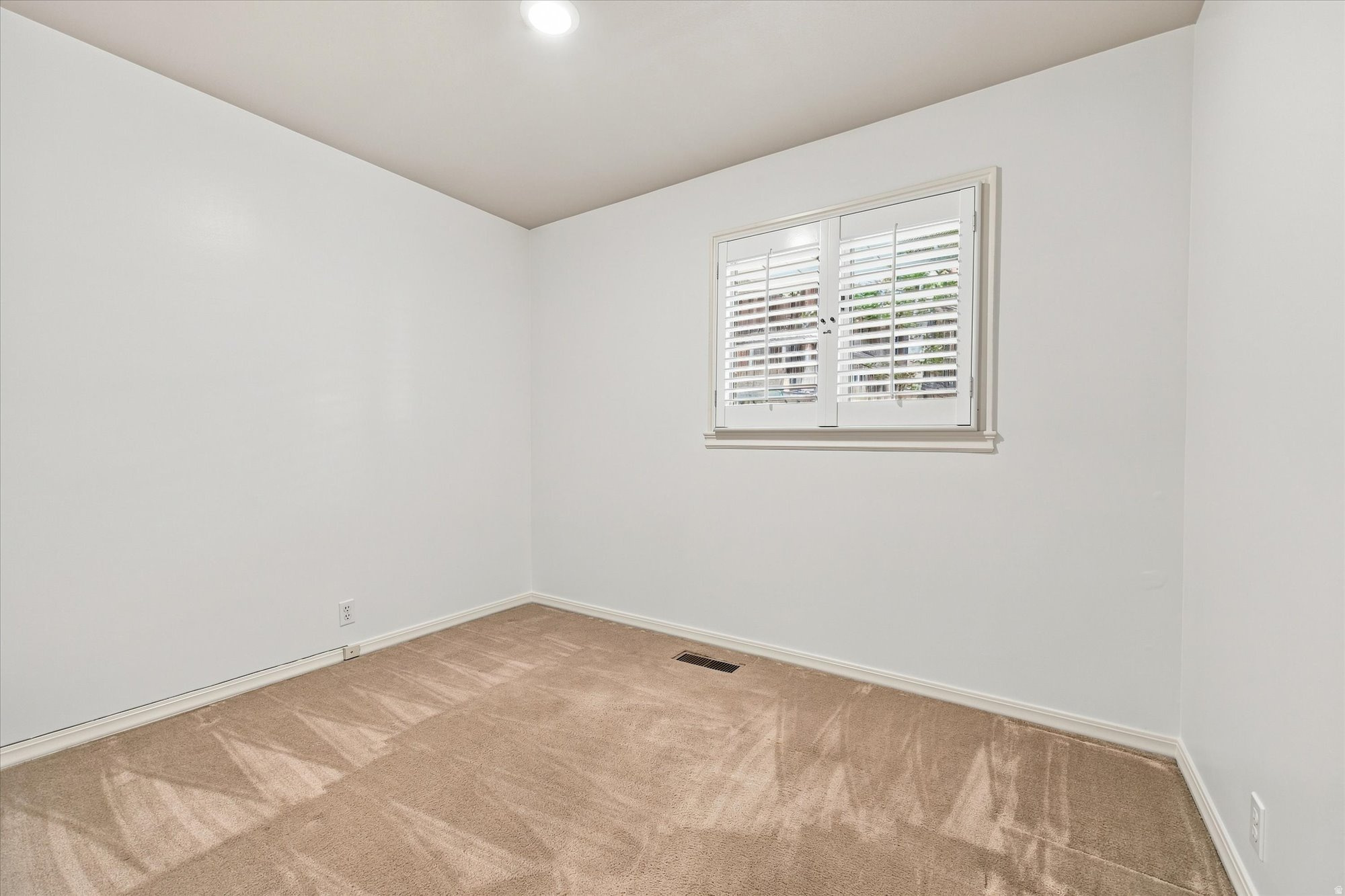 Empty carpeted room with white plantation-shuttered window and recessed ceiling light.