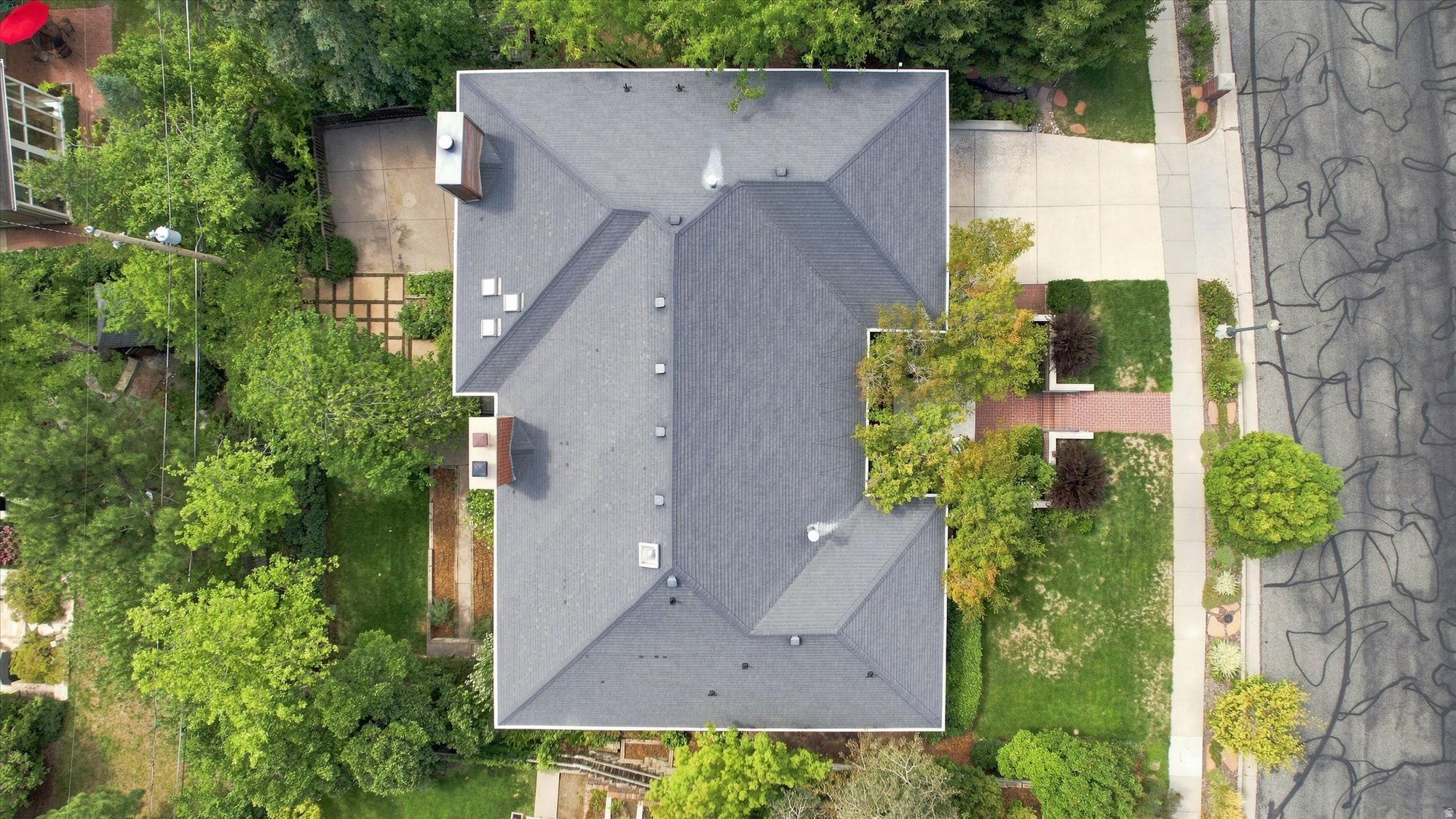 Aerial view of a gray-roofed home surrounded by mature trees, landscaped yards, and a sidewalk.
