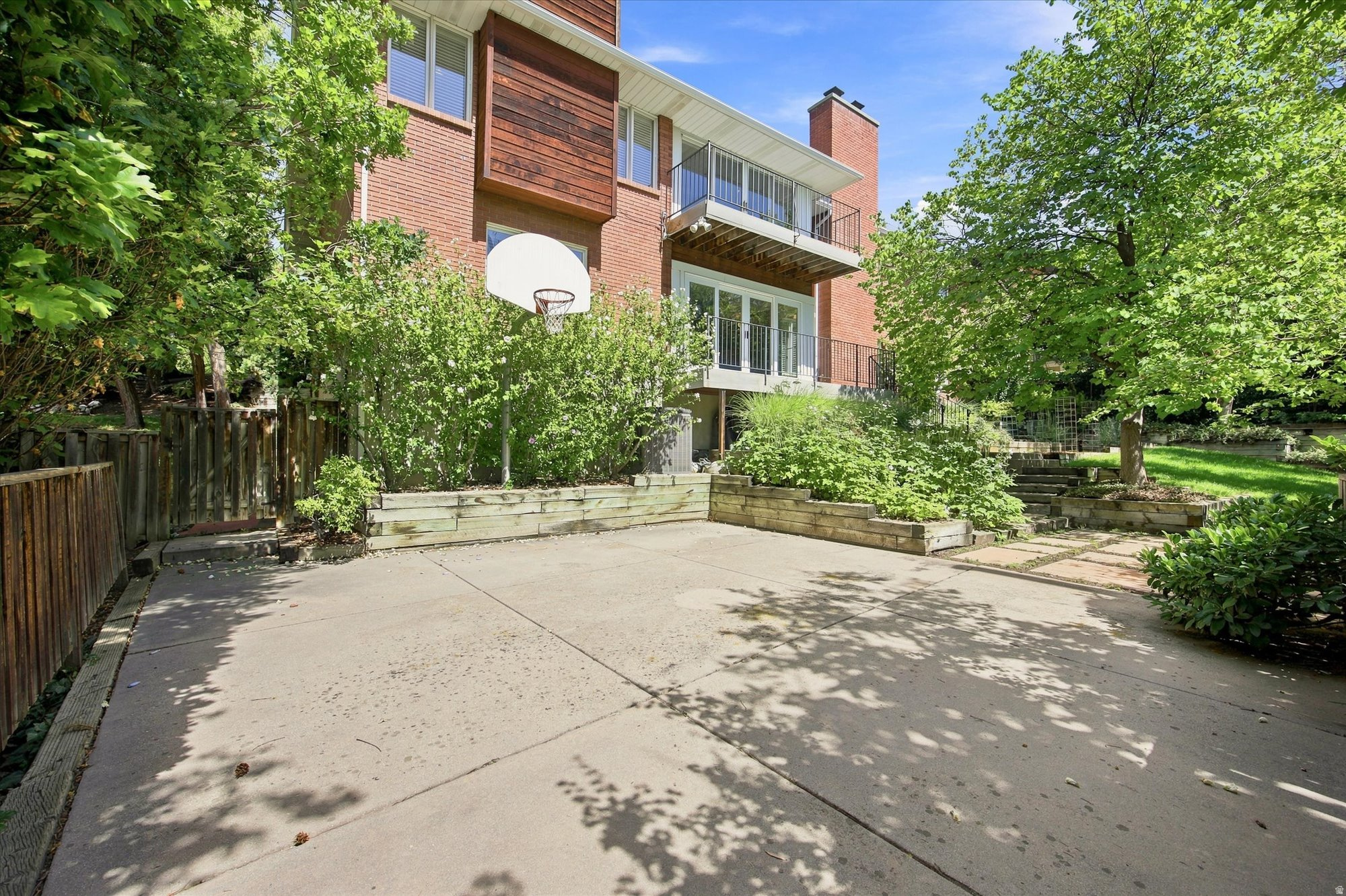 Brick home’s rear patio with balconies, raised garden beds, mature trees, and a basketball hoop.