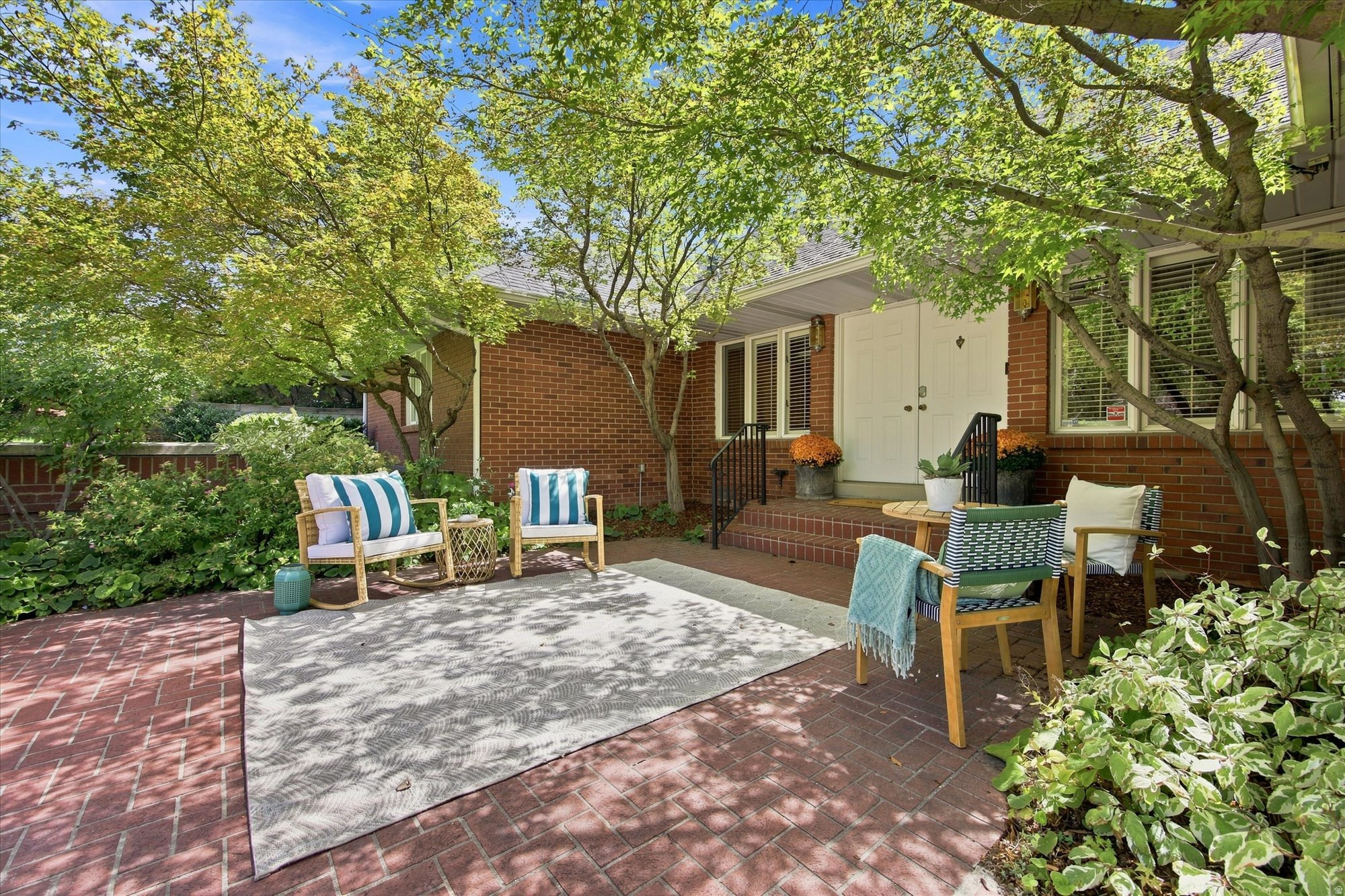 Brick patio with seating beneath mature trees outside a red-brick home's front entrance.
