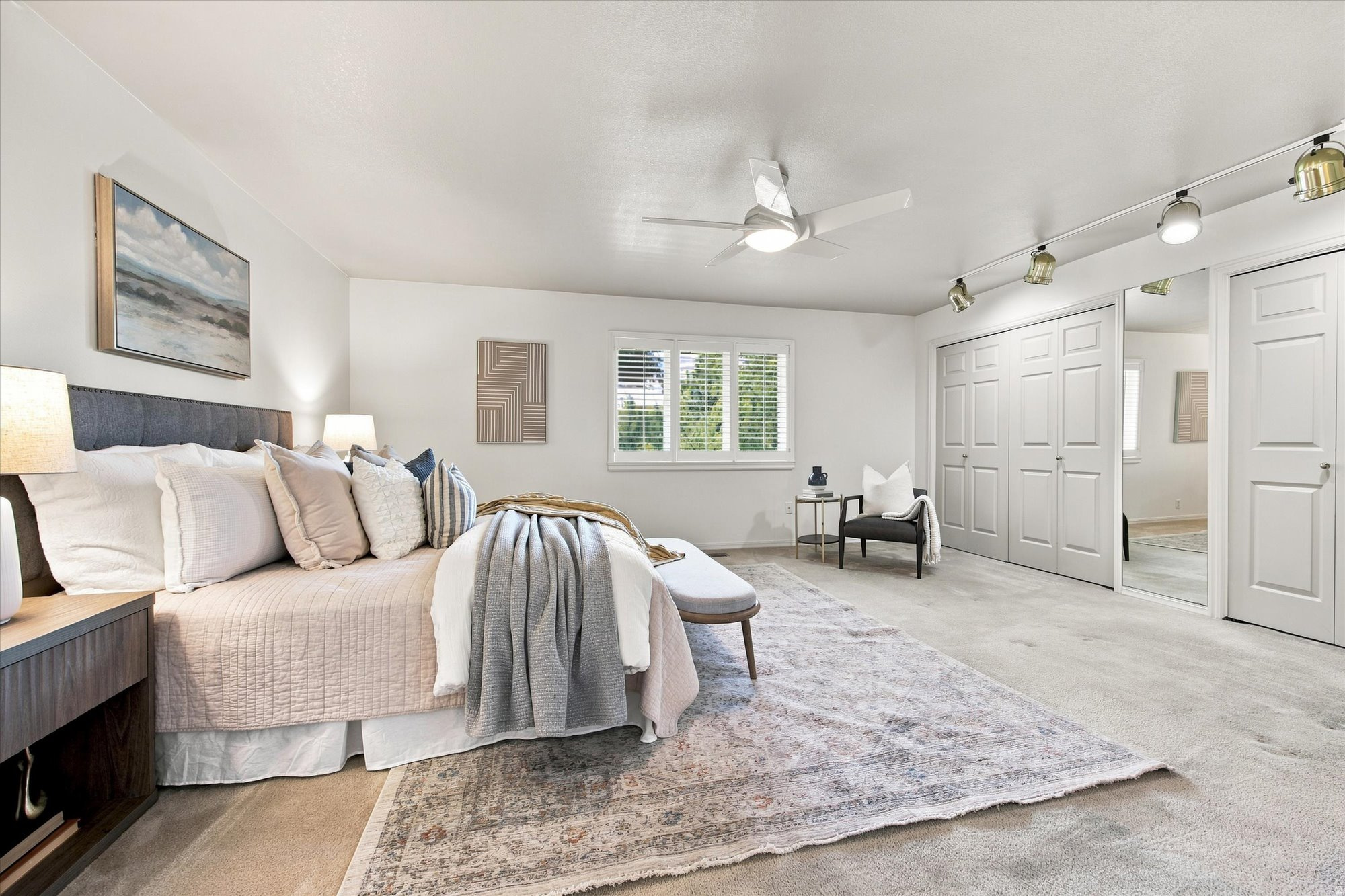Staged bedroom with a large bed, plantation-shuttered window, mirrored closet doors, and pale patterned rug.
