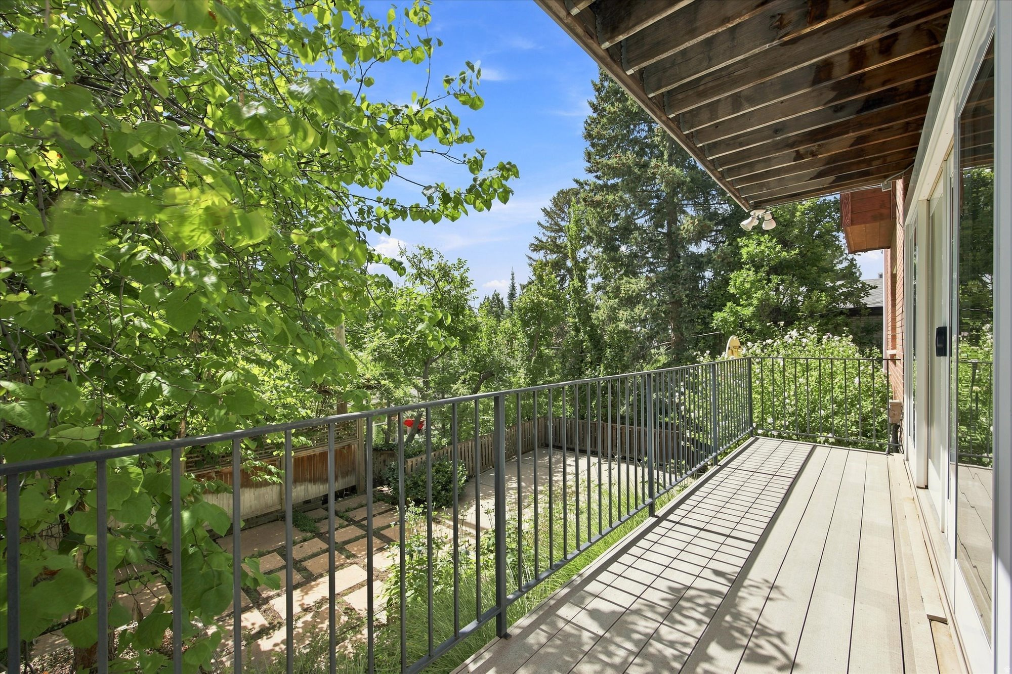 Covered wooden deck with metal railing overlooking a leafy garden beside sliding glass doors.