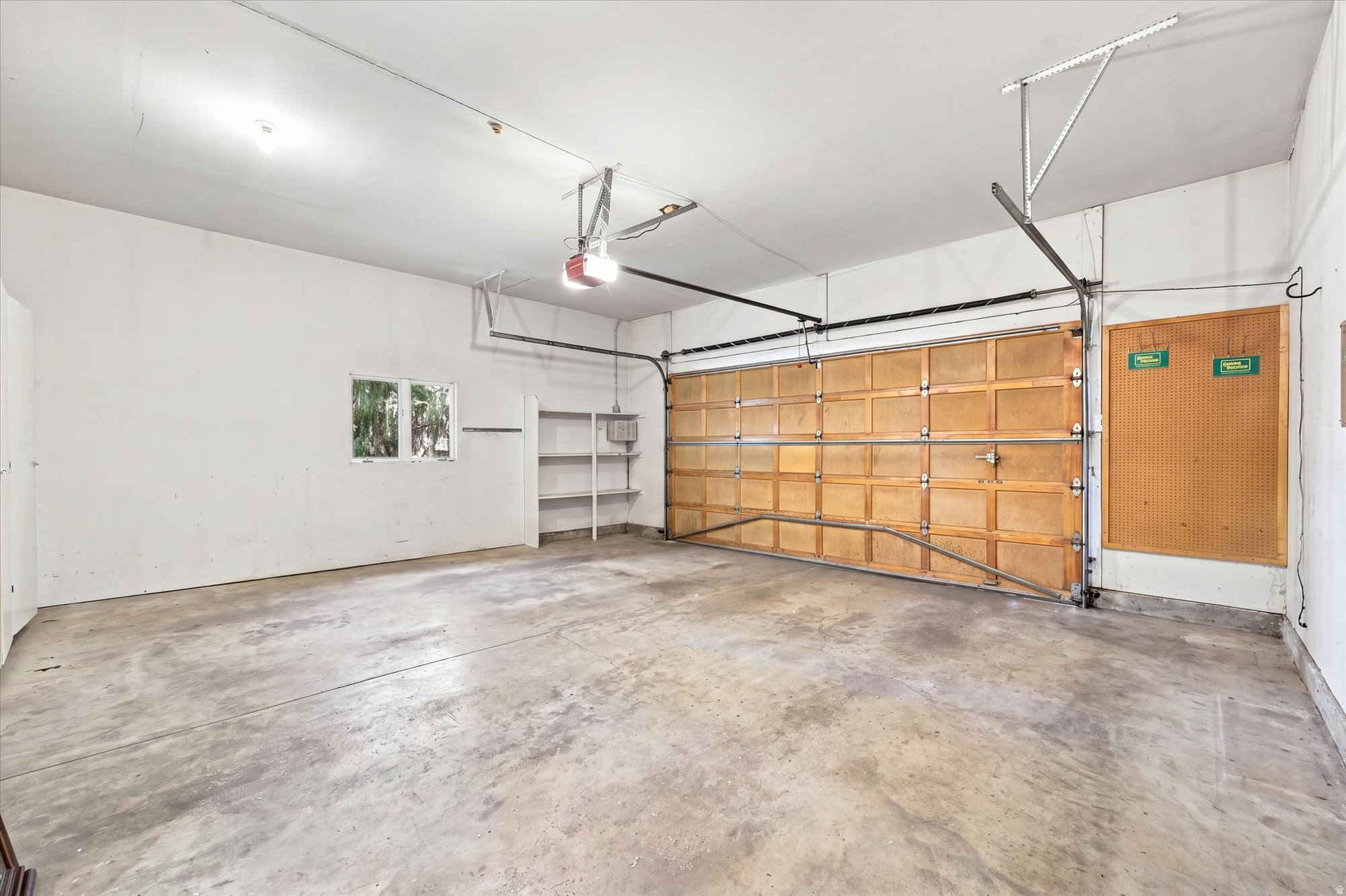 Empty garage with a wide wooden door, concrete floor, shelving, and pegboard.