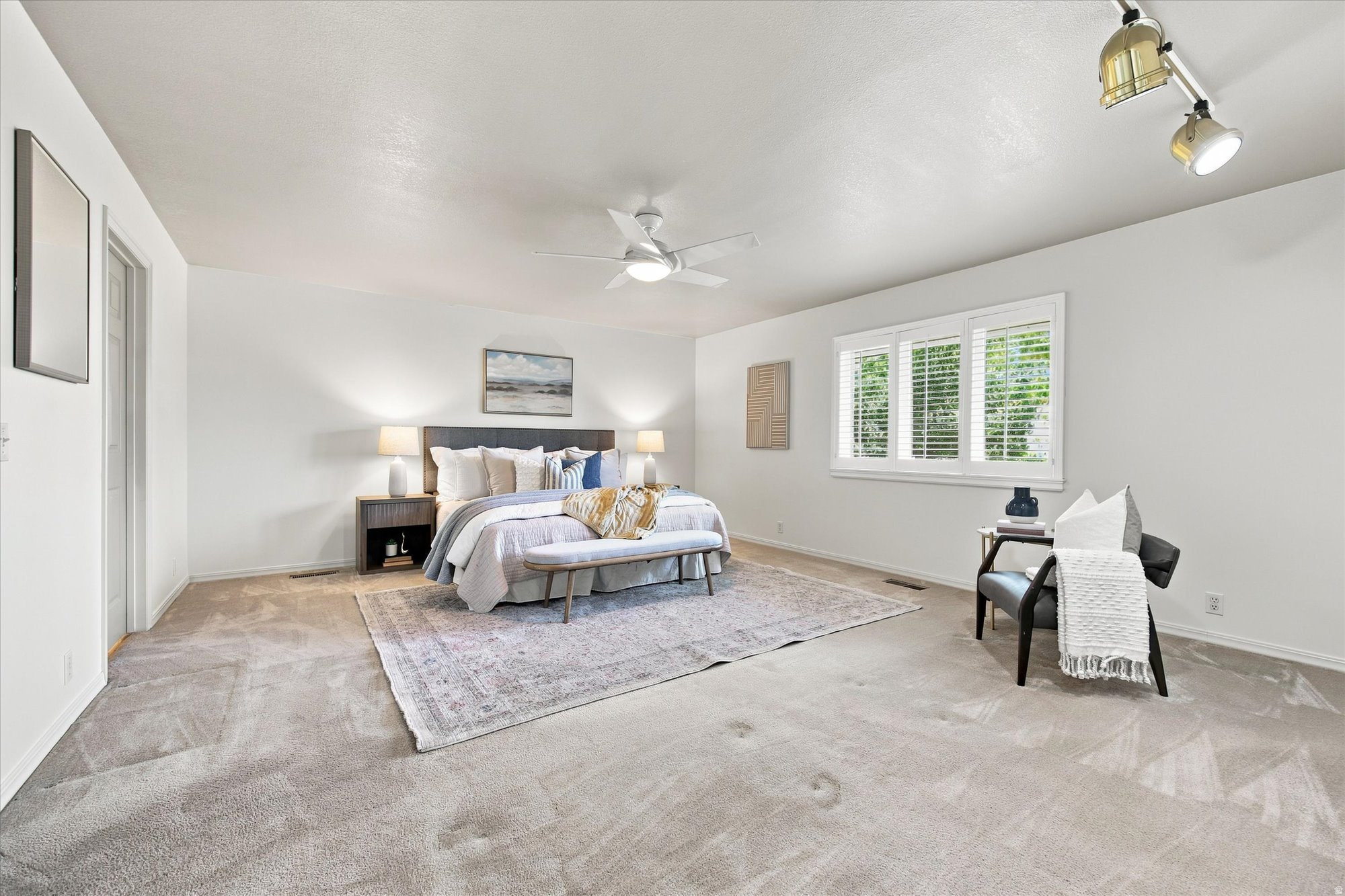 Staged bedroom with a large bed, plantation shutters, and a black armchair with a white throw.