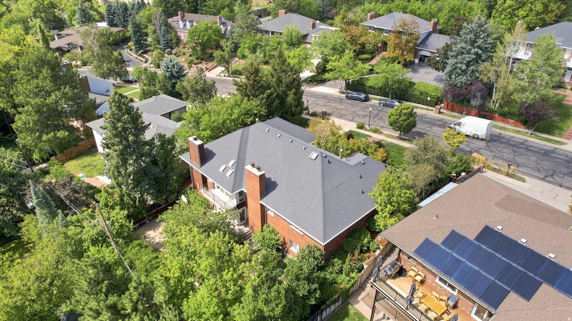 Aerial view of a red-brick home with a broad gray roof among mature trees and neighboring houses.
