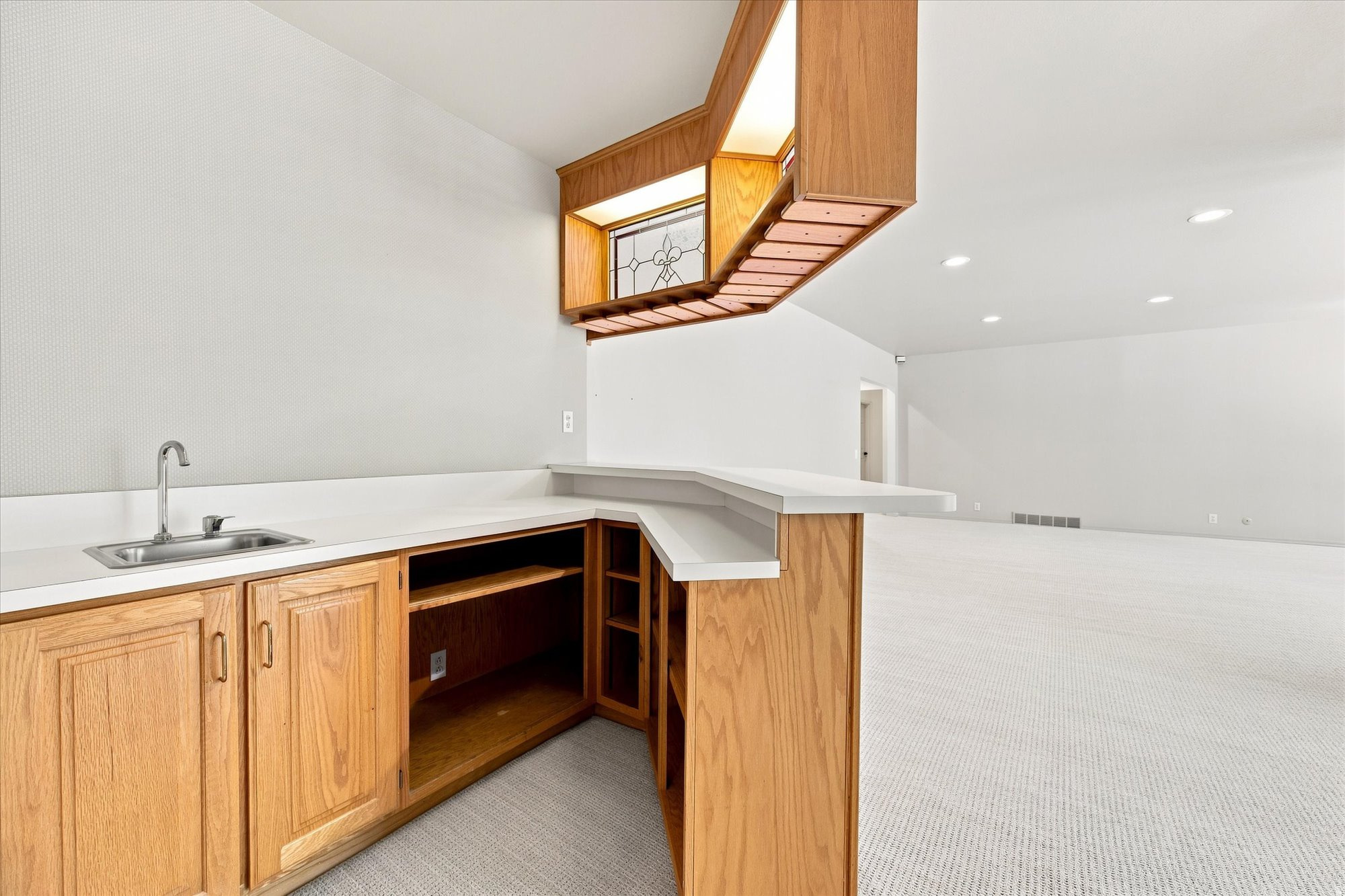 Empty wet bar with honey-colored wood cabinets, stainless steel sink, white counters, and a stained-glass window.