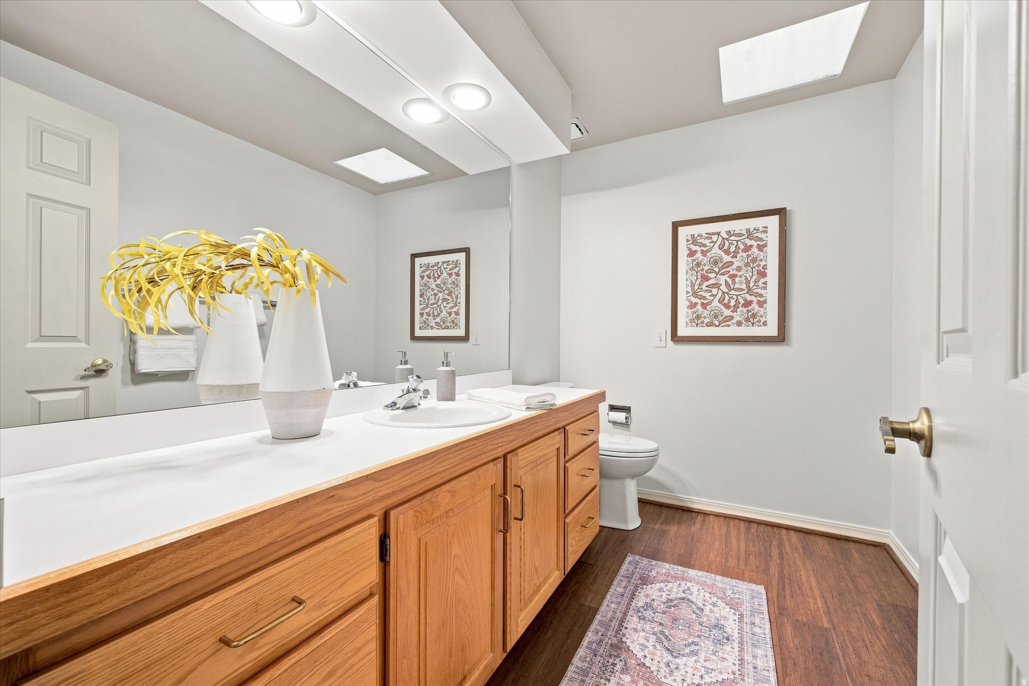 Bathroom with a long oak vanity, skylights, and framed floral artwork.