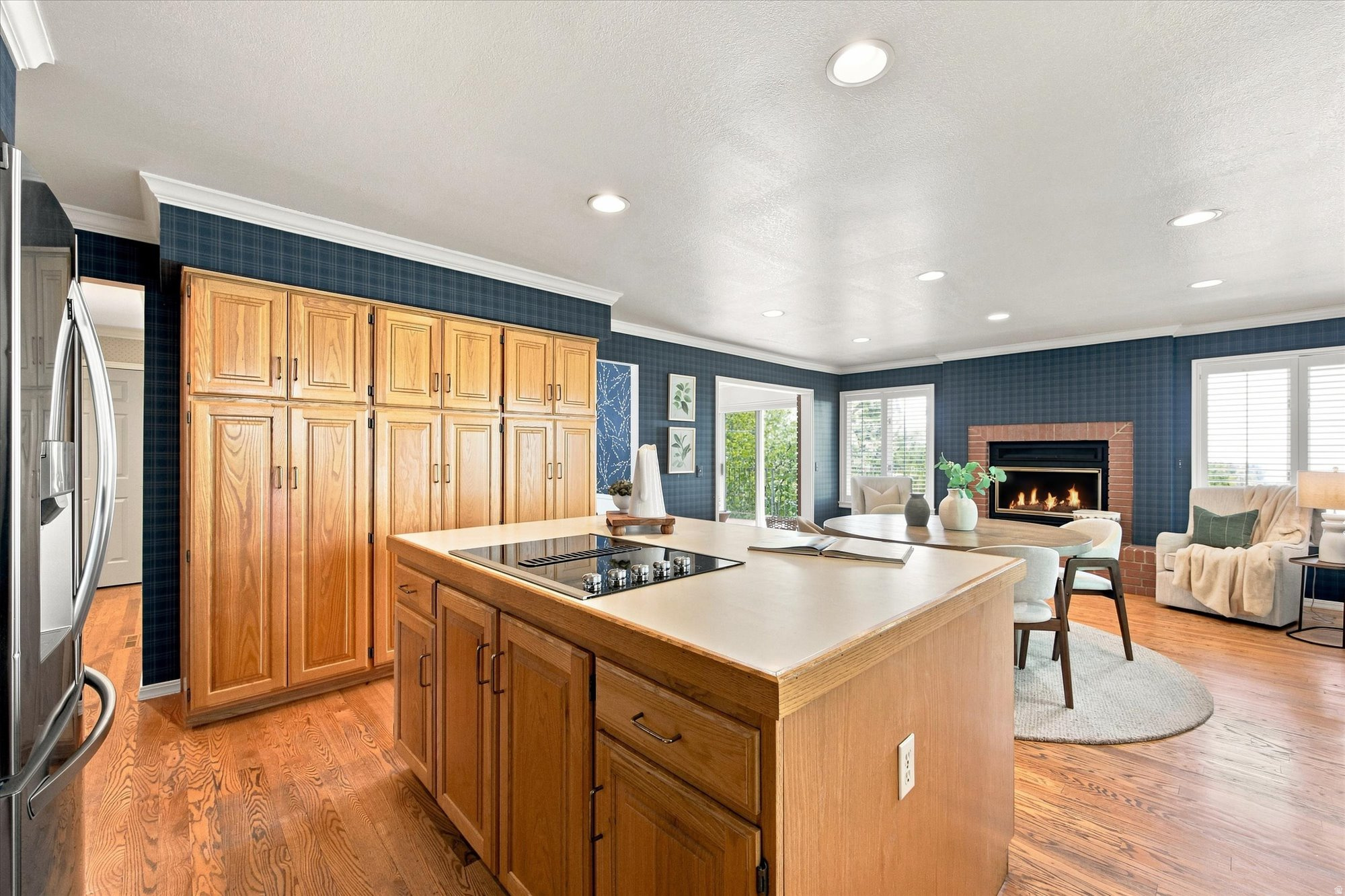 Kitchen with a large wood island and cooktop opening to a sitting area with a lit brick fireplace.