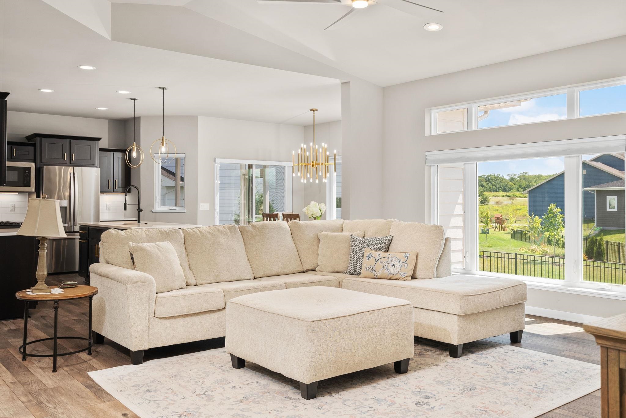 Open-concept living room with a cream sectional and ottoman, dark kitchen, and wide windows overlooking a yard.